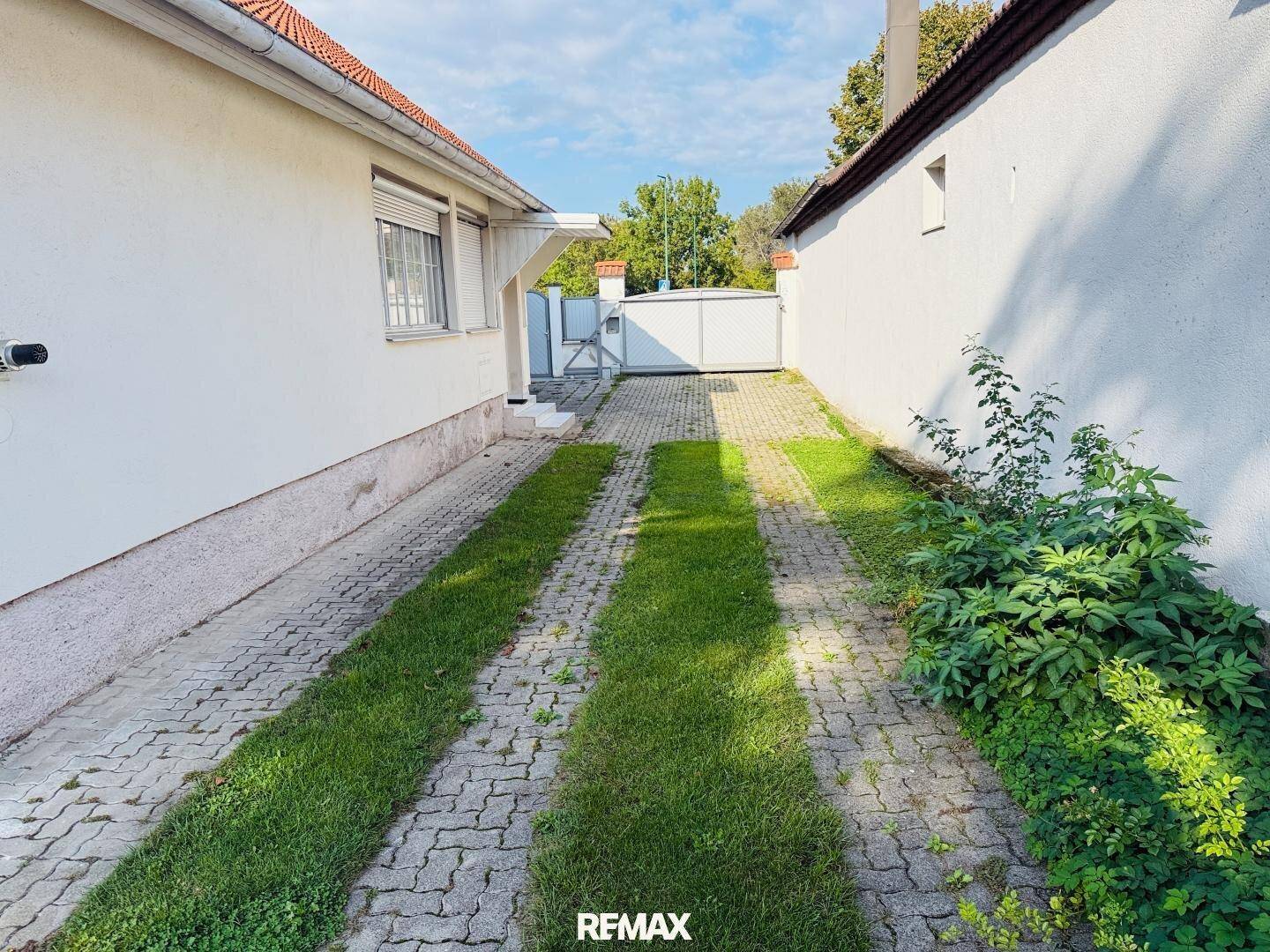 Schmaler gepflasterter Weg zwischen zwei Gebäuden mit Grasstreifen und Zaun im Hintergrund.