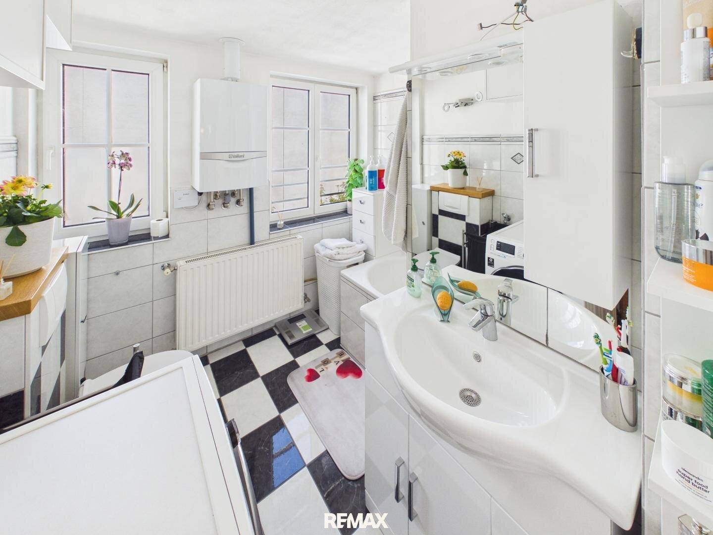 Helles Badezimmer mit Waschbecken, Dusche, Fenster und schwarz-weiß kariertem Fliesenboden.