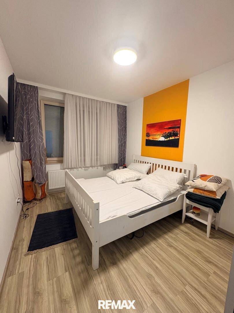Schlafzimmer mit weißem Bett, Laminatboden und einer farbigen Akzentwand, ausgestattet mit einem Fenster.