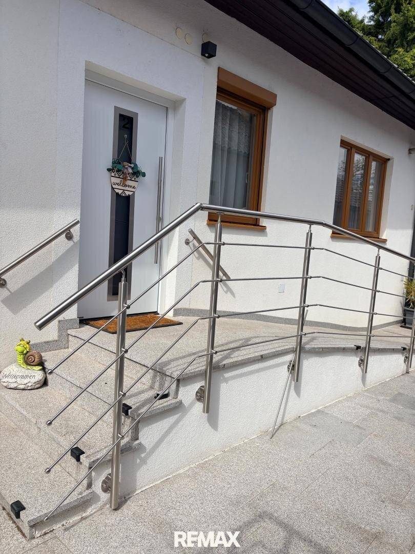 Hauseingang mit Treppe und Rampe, gesichert durch ein Edelstahlgeländer, umgeben von einer gepflegten Außenanlage.