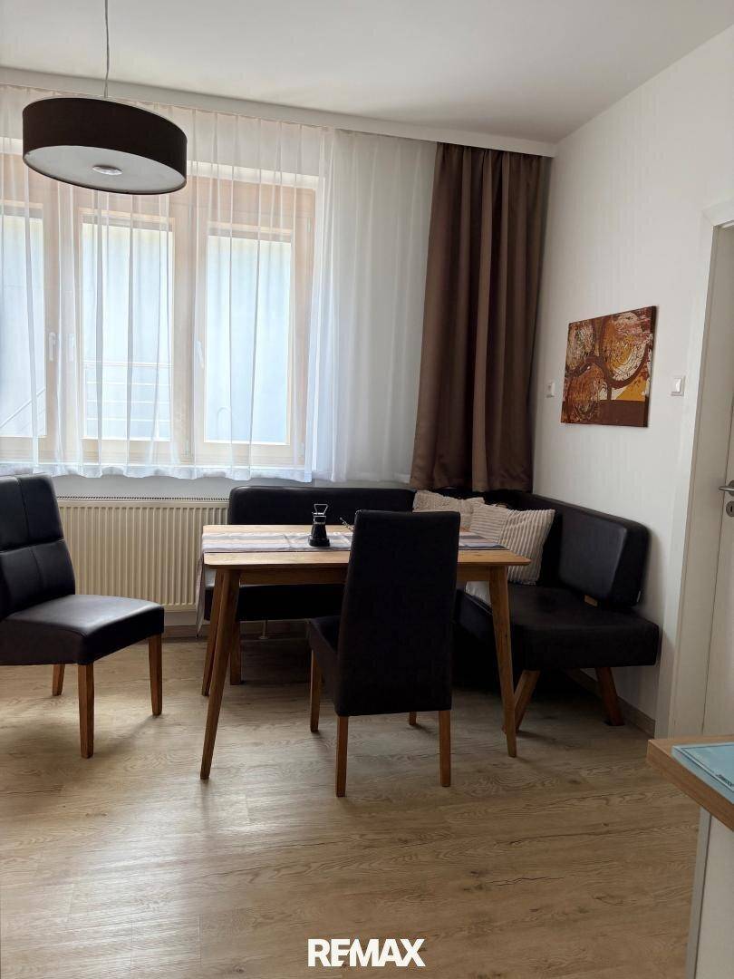 Heller Essbereich mit Holztisch, dunklen Stühlen und Sitzbank, umgeben von großen Fenstern mit Vorhängen.