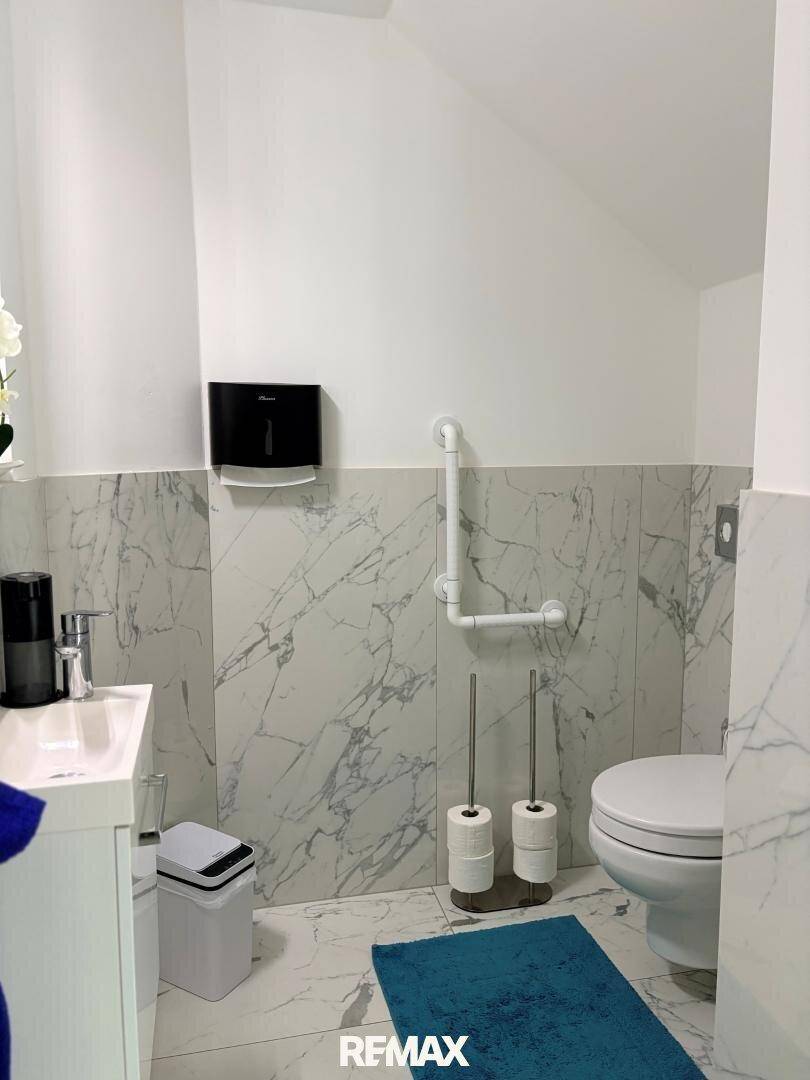Modernes Gäste-WC mit Wand-WC, kleinem Waschbecken und Marmoroptik-Fliesen, ausgestattet mit Haltegriffen.