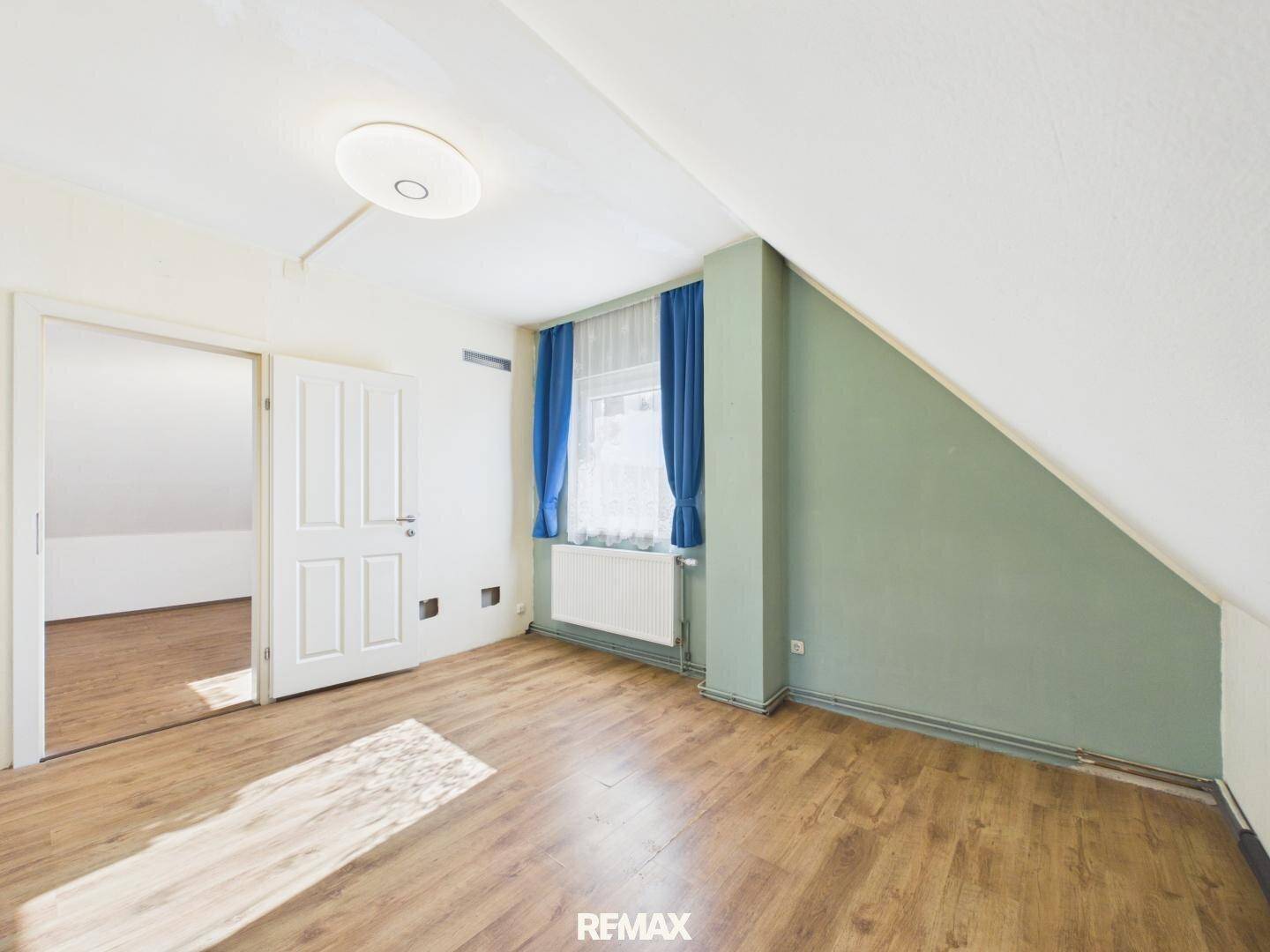 Dachgeschosszimmer mit Laminatboden, grüner Akzentwand und Fenster mit blauen Vorhängen.