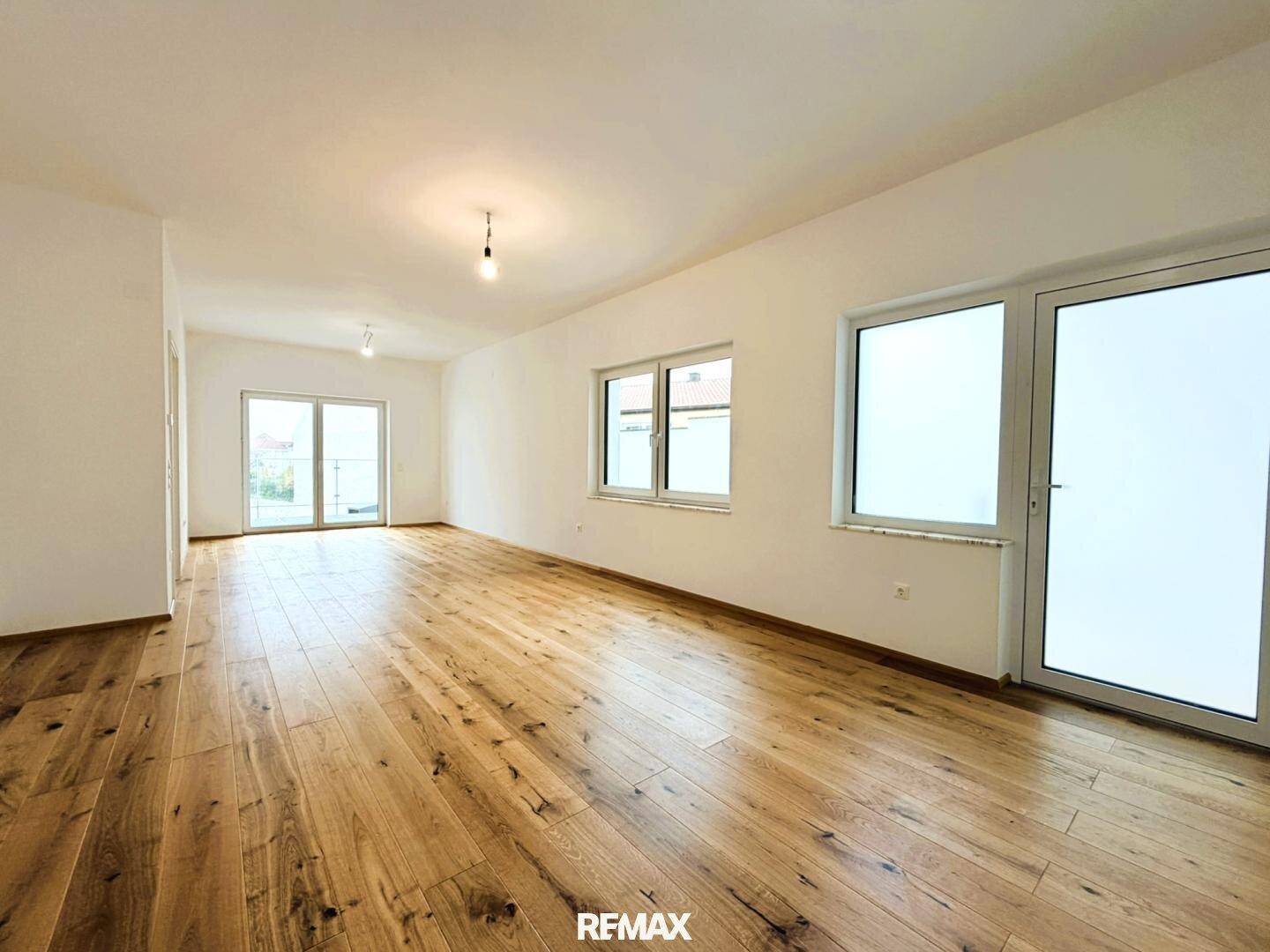 Heller, leerer Wohnraum mit Holzboden und großen Fenstern, die viel Tageslicht hereinlassen.