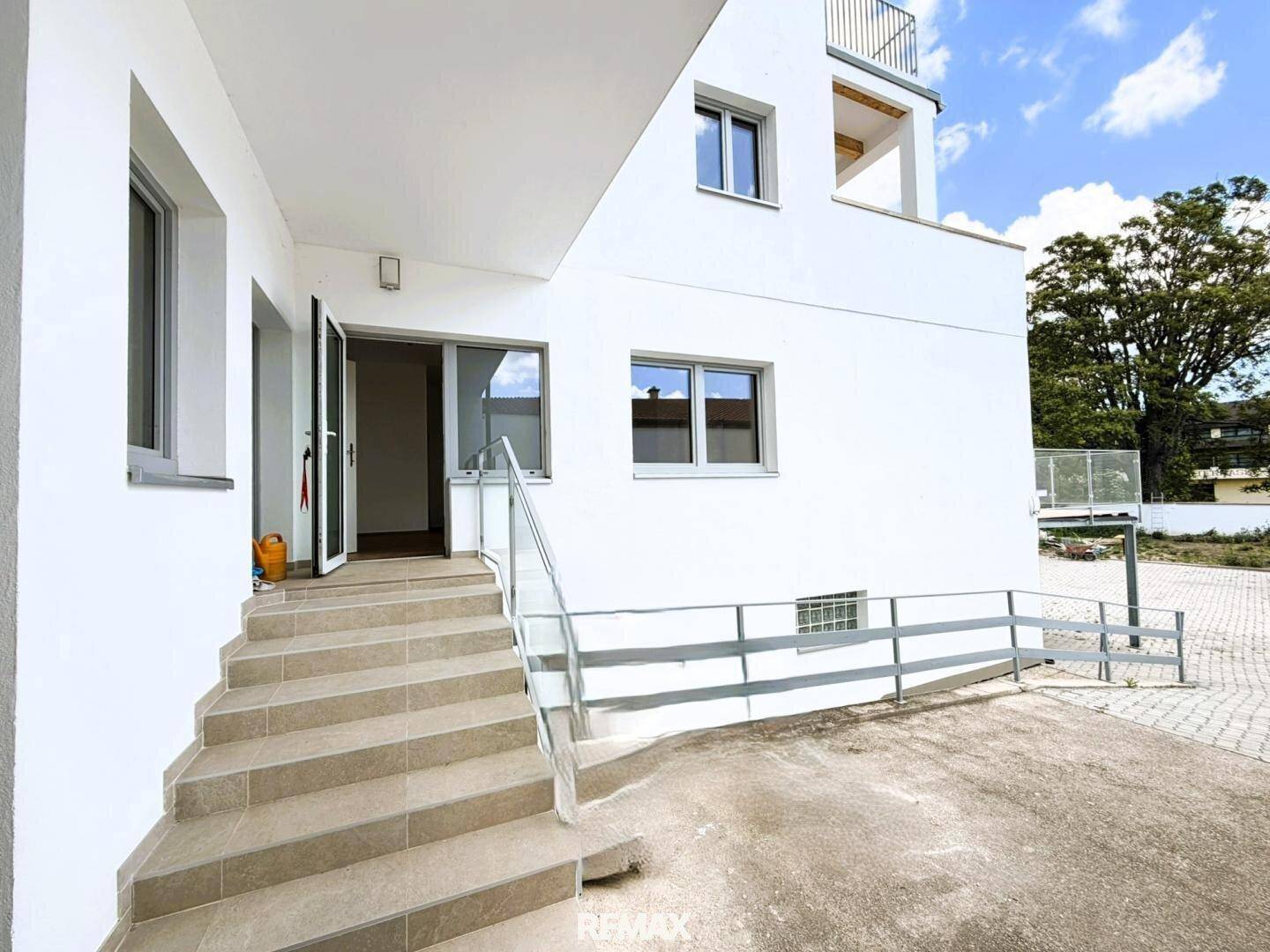 Moderne weiße Gebäudefassade mit Treppe und Rampe zum Eingangsbereich unter blauem Himmel.