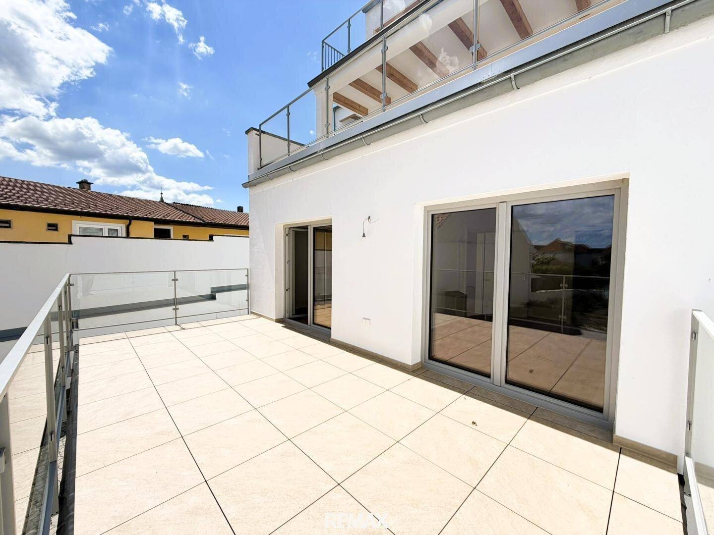 Geräumige Dachterrasse mit hellen Fliesen, Glasgeländer und weitem Blick unter blauem Himmel.