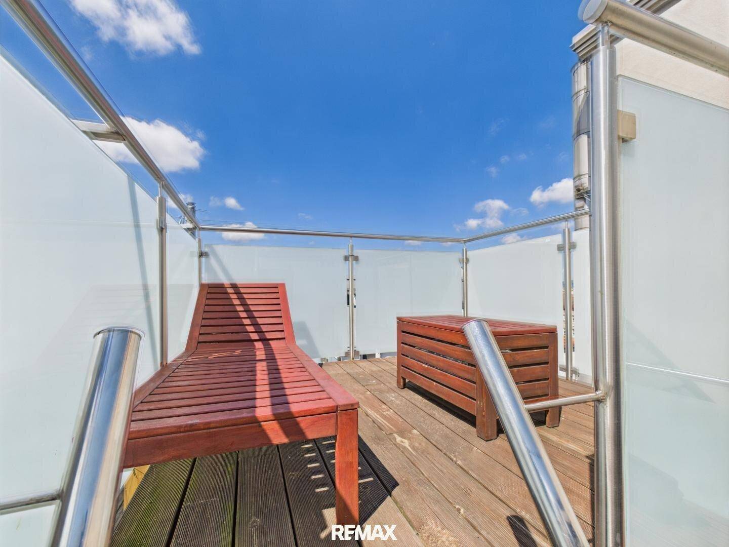 Sonniger Balkon mit Holzliege und Glasgeländer, ideal zum Entspannen im Freien.