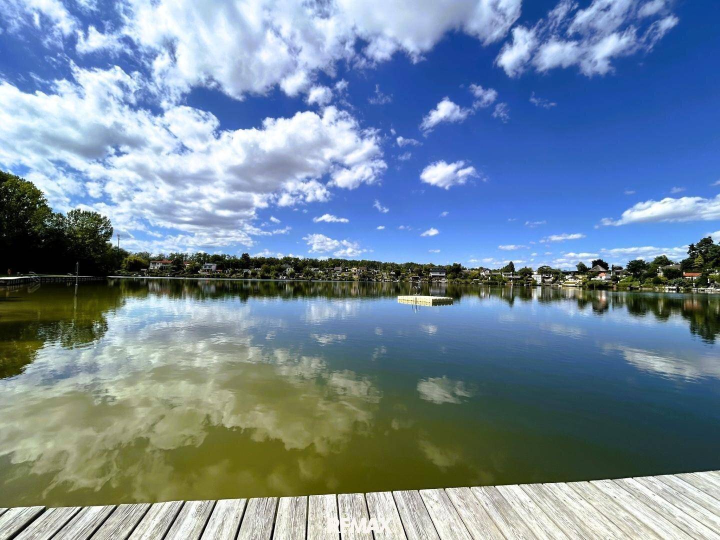 Malerischer See mit Holzanleger und Häusern am Ufer unter einem bewölkten Himmel.