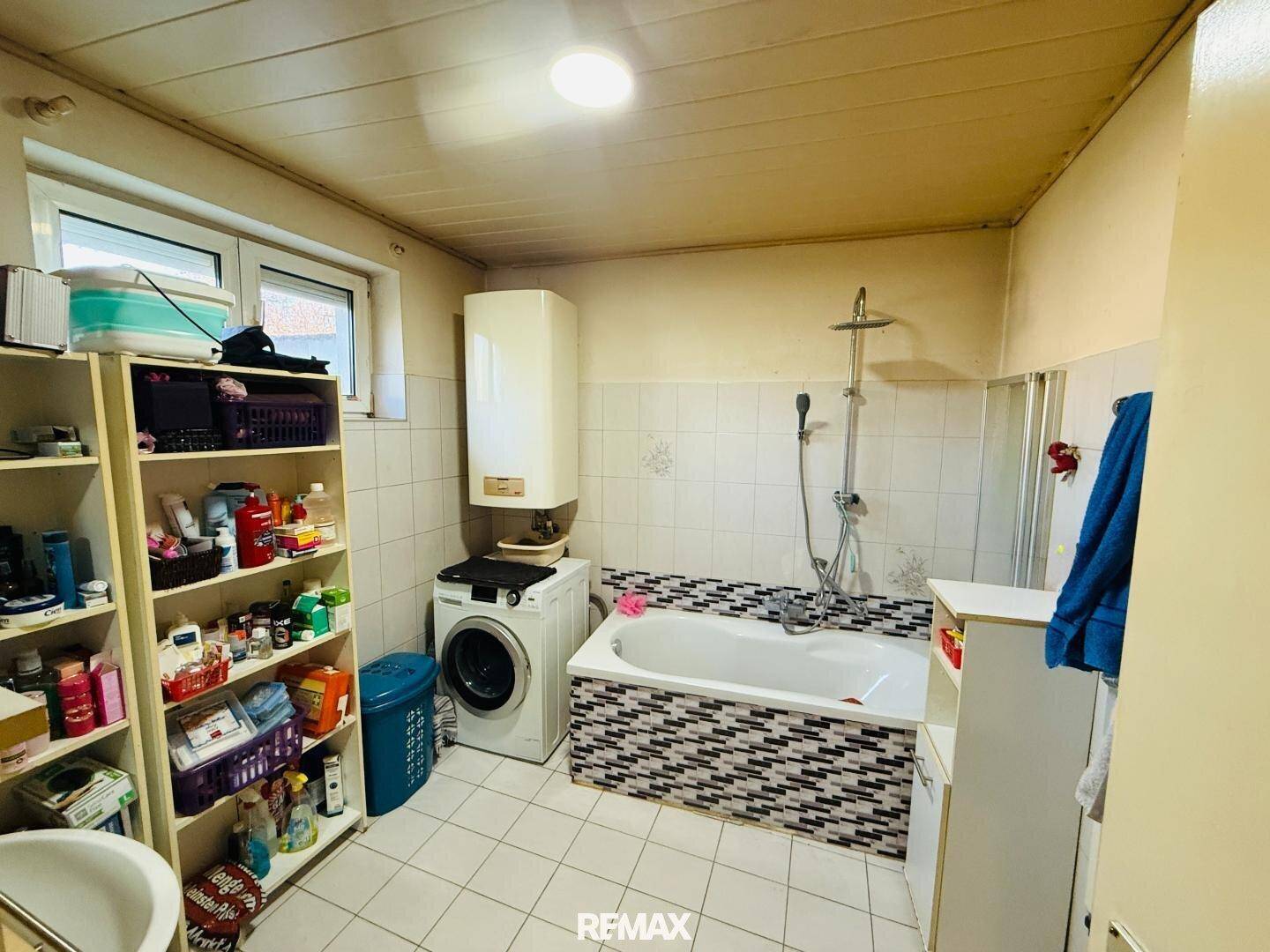 Badezimmer mit Badewanne, Dusche und Waschmaschine, hell gefliest und funktional ausgestattet.