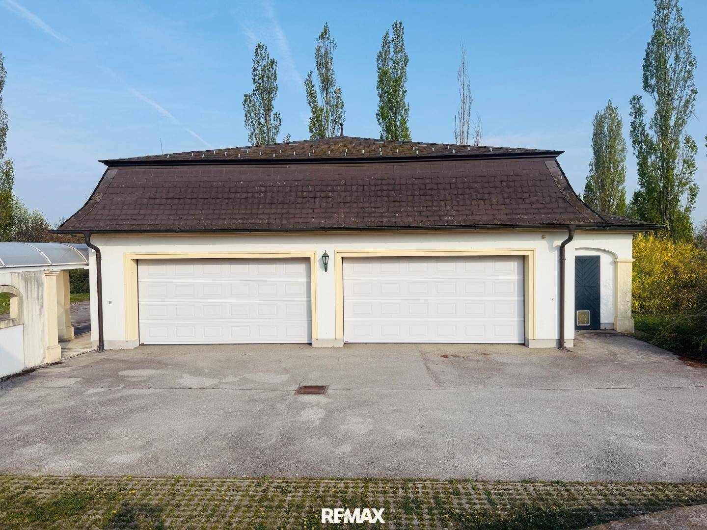 Geräumiges Garagengebäude mit zwei weißen Toren und einem Mansarddach, umgeben von hohen Bäumen.