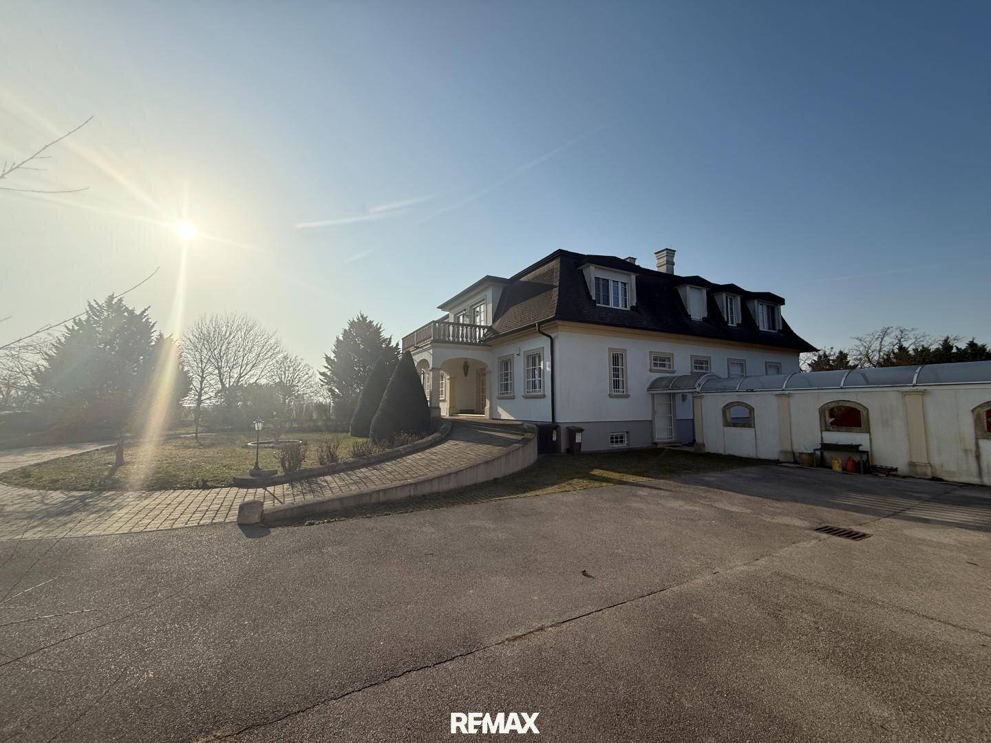Seitenansicht der Villa mit weitläufiger Auffahrt und angrenzendem Garagengebäude bei Sonnenschein.