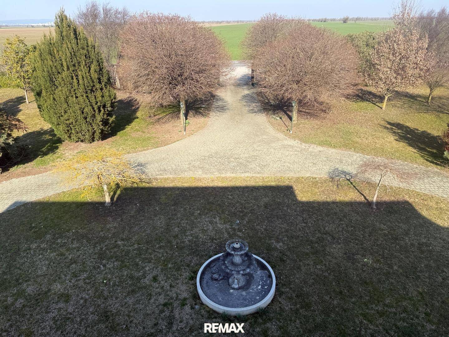 Panoramablick von oben auf den Garten mit Brunnen und die weite, grüne Landschaft im Hintergrund.