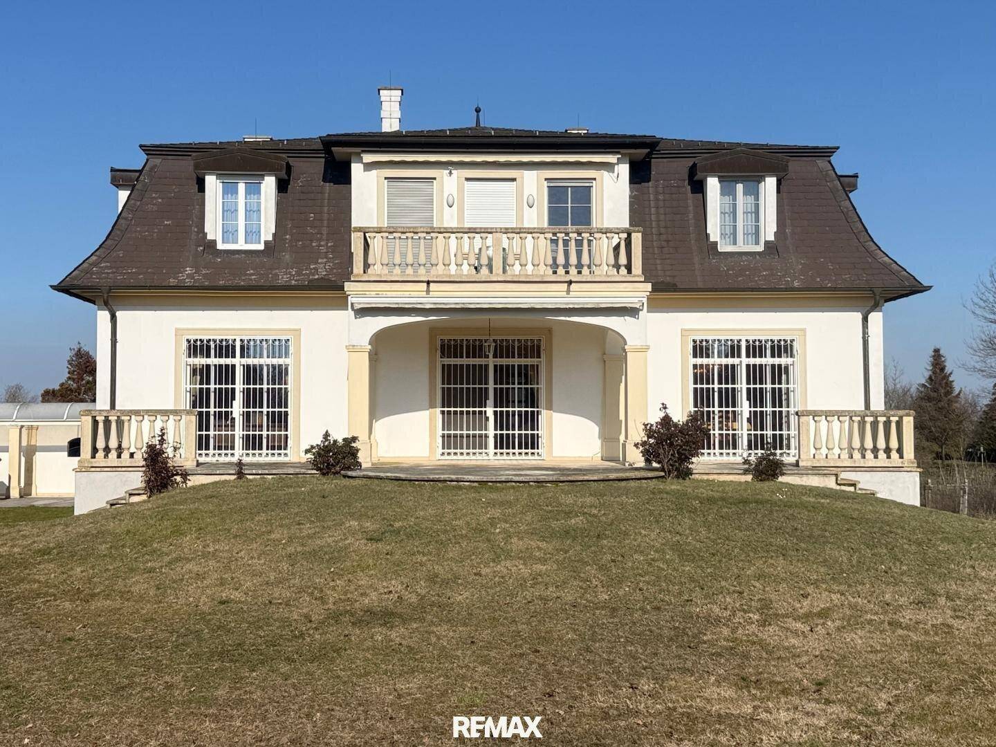 Elegante Villa mit Mansarddach, Balkon und großen Fenstern auf einem Hügel.