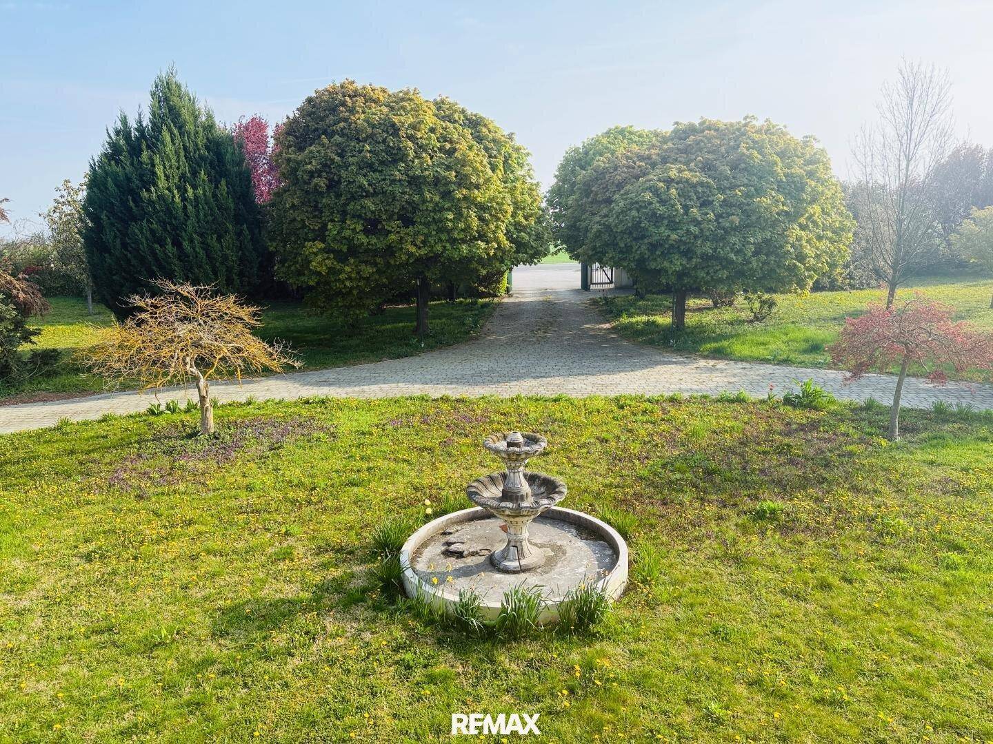 Idyllischer Gartenbereich mit einem klassischen Brunnen und altem Baumbestand unter strahlendem Himmel.