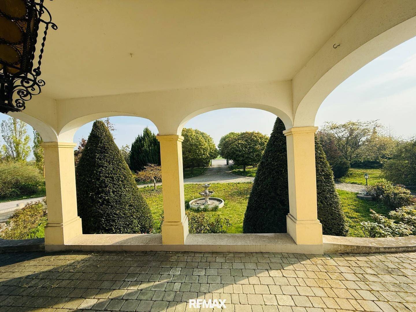 Blick vom überdachten Eingangsbereich auf den weitläufigen Garten mit Brunnen und gepflegten Bäumen.