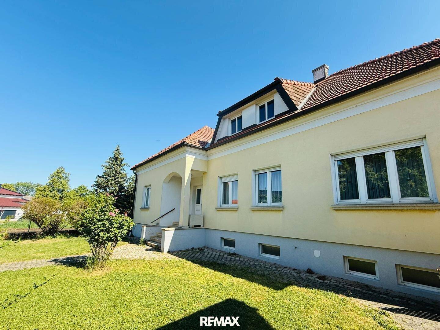 Seitenansicht des Hauses mit gelber Fassade, roten Dachziegeln und einem sonnigen Vorgarten.