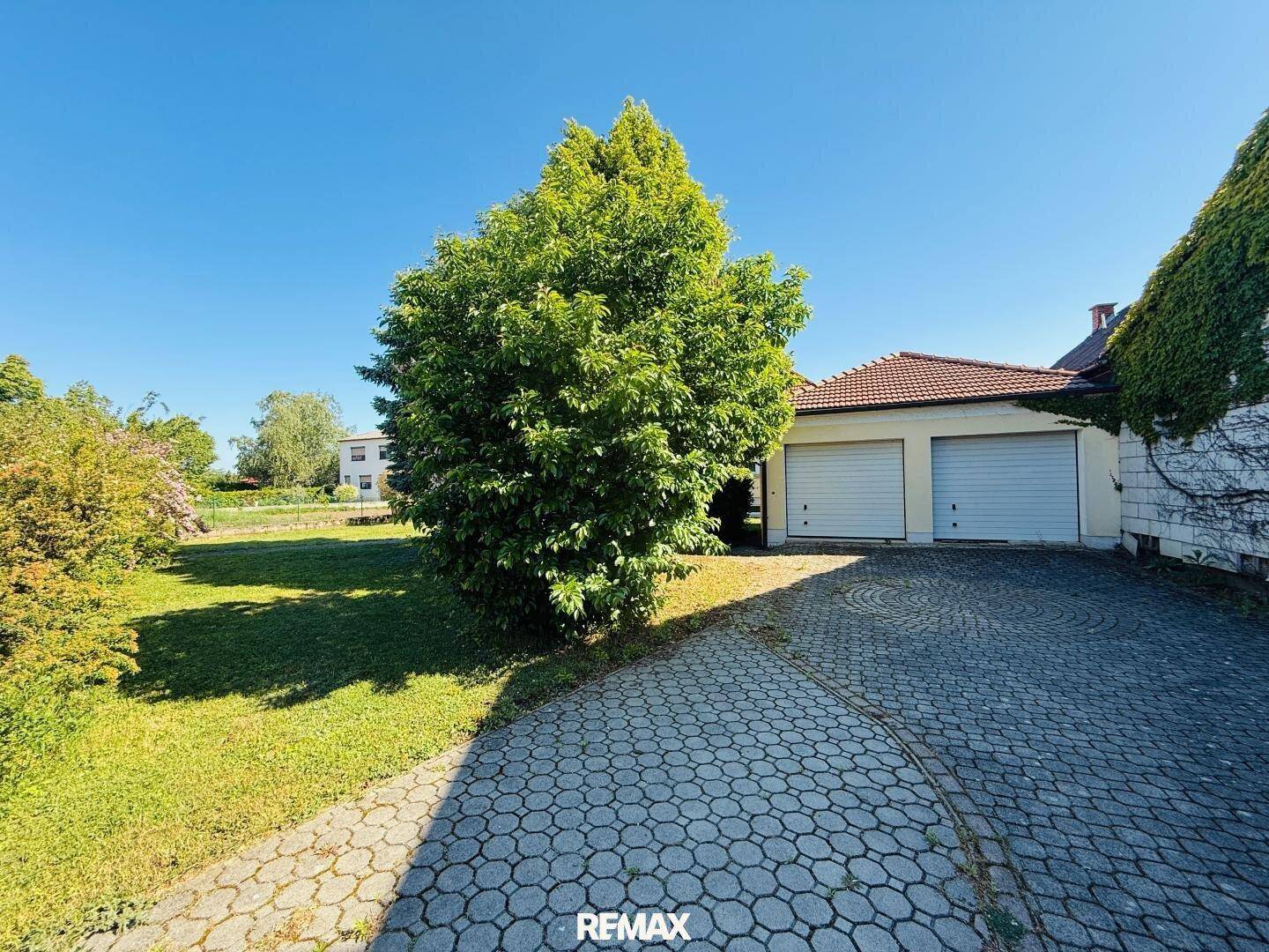 Doppelgarage mit gepflasterter Auffahrt und einem großen Baum im sonnigen Garten.