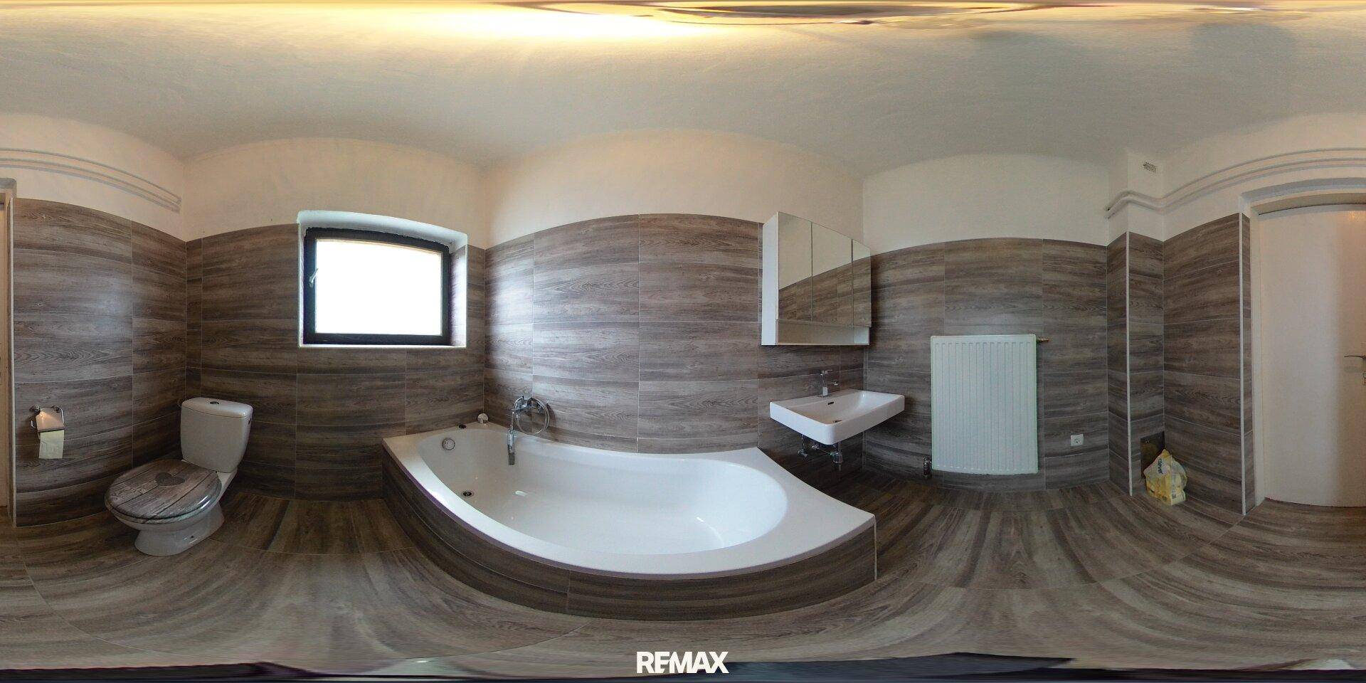 Badezimmer mit Eckbadewanne, Waschbecken, WC, Spiegelschrank und holzähnlichen Fliesen.