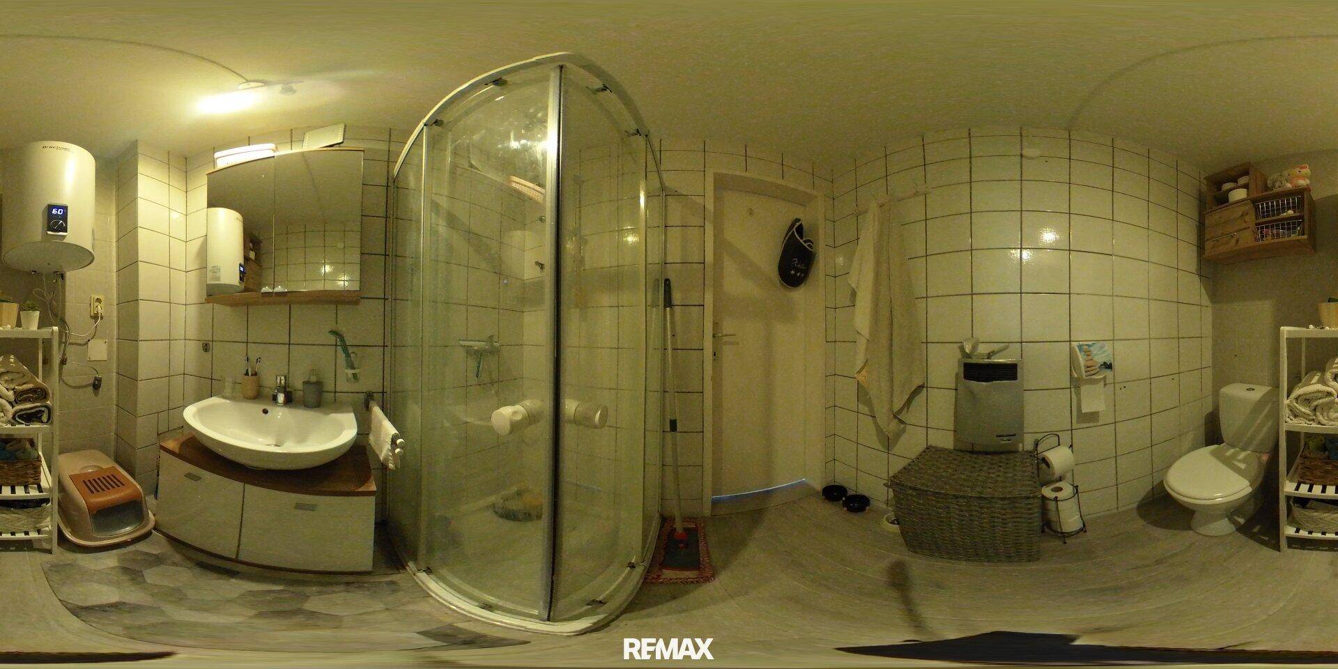 Panoramaansicht des Badezimmers mit Duschkabine, Toilette und hellgrauen Fliesen an den Wänden.