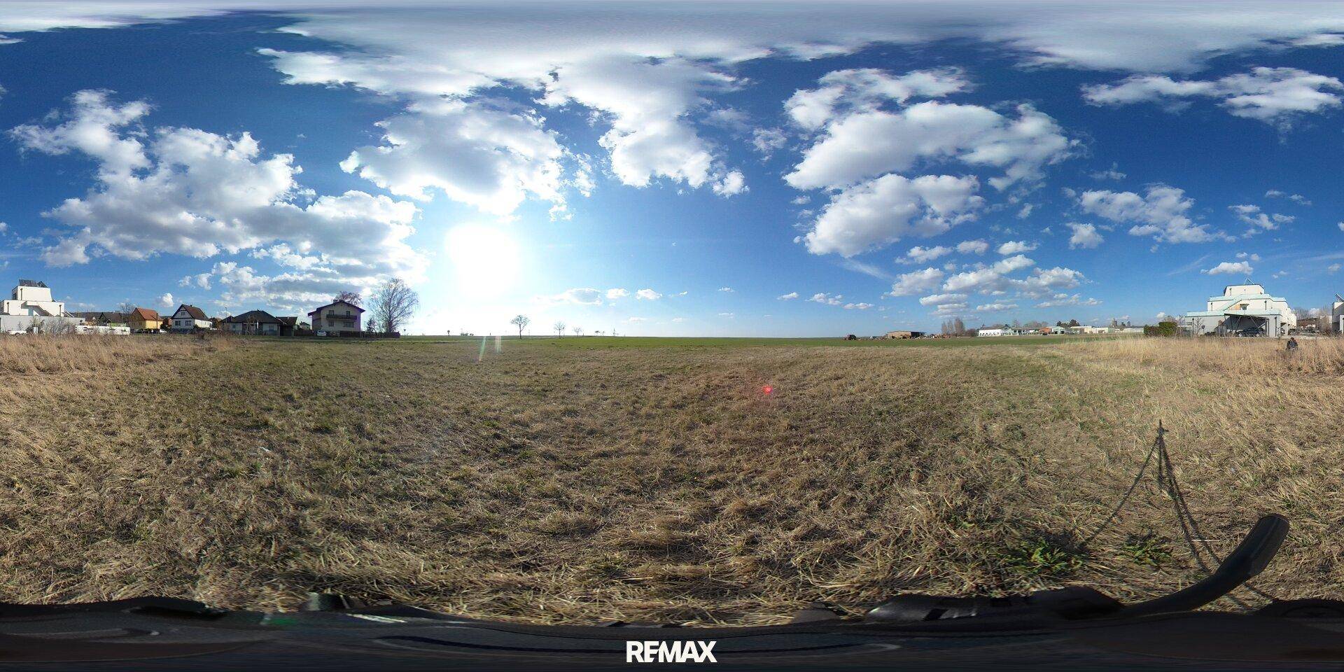 Panoramaaufnahme eines weiten Feldes mit blauem Himmel und vereinzelten Gebäuden in der Ferne.