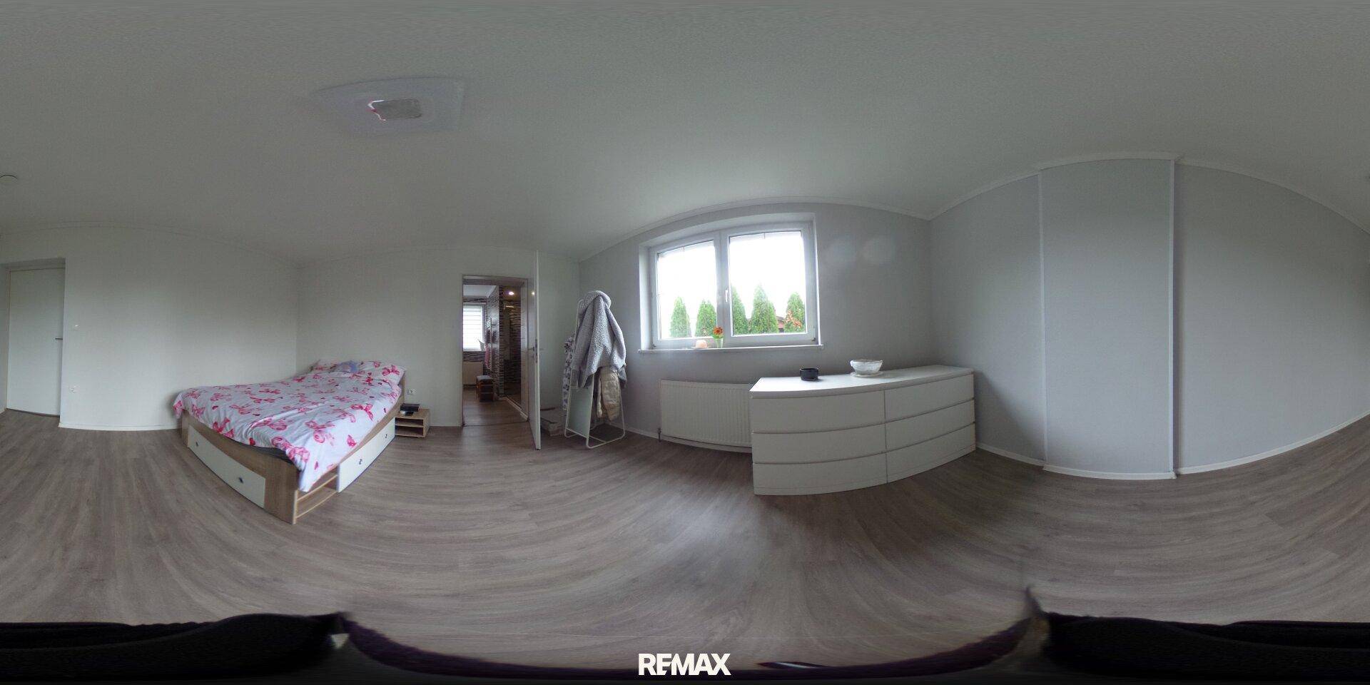 Helles Schlafzimmer mit großem Fenster, Laminatboden und einem modernen Bett mit floralem Muster.