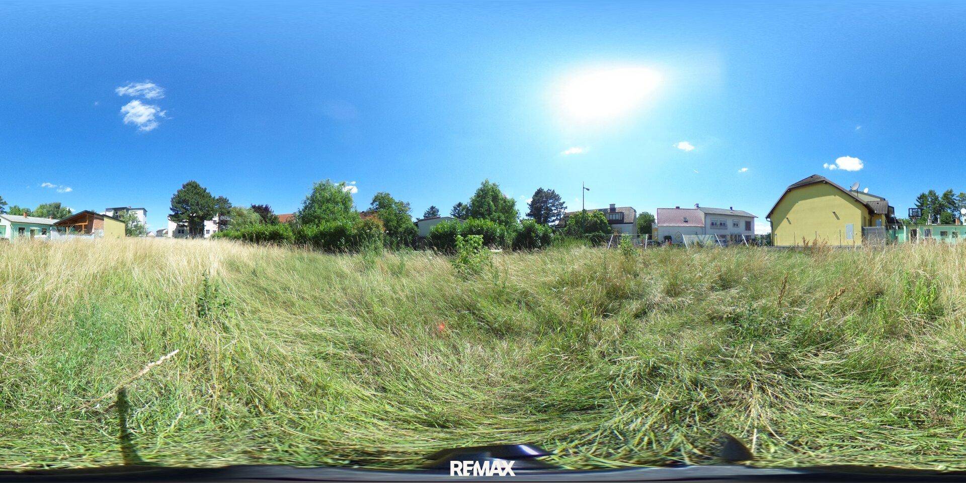 Panoramablick auf ein sonniges Grundstück mit hohem Gras und umliegender Wohnbebauung.