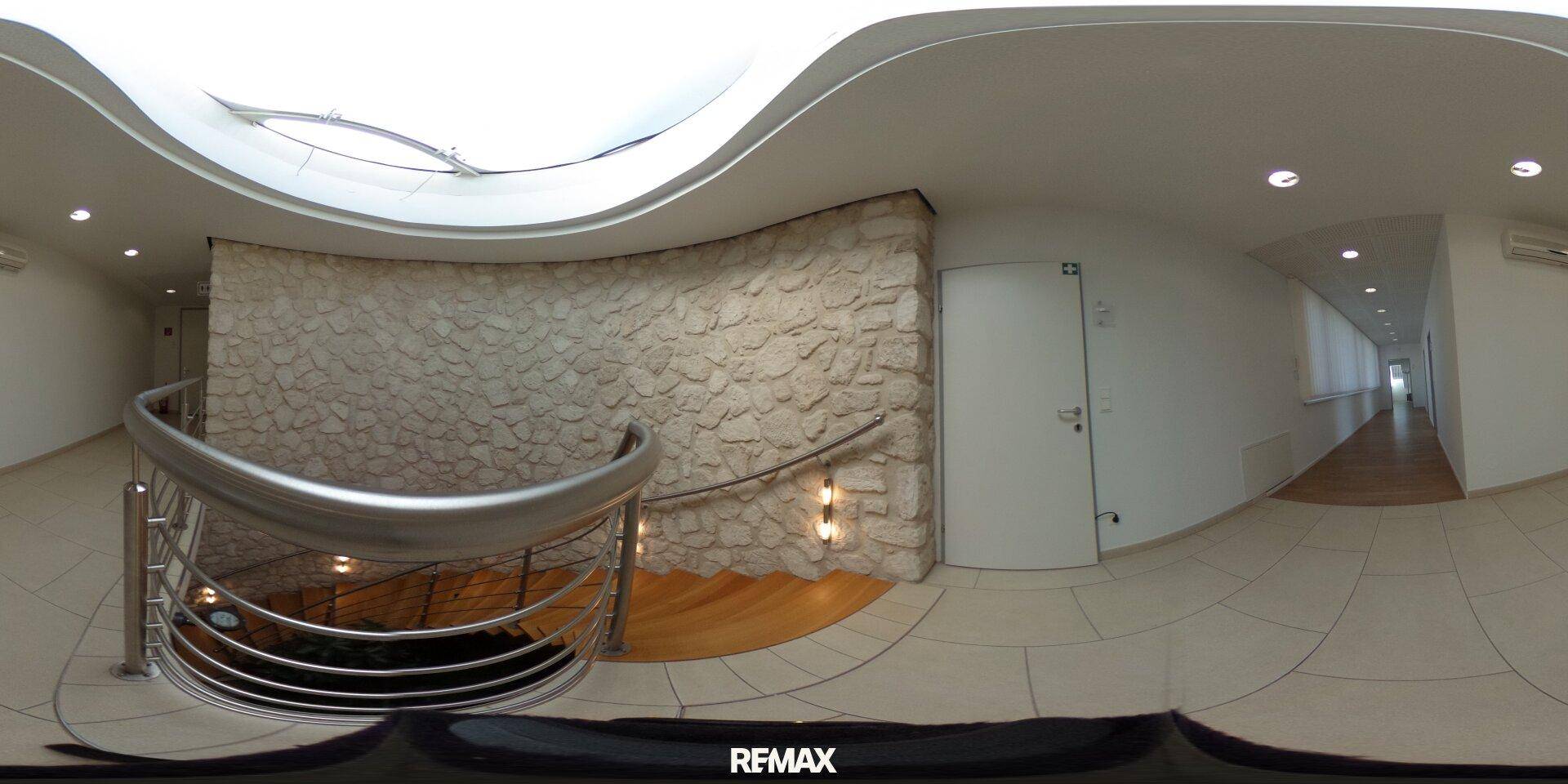 Panoramaansicht eines Flurs mit Steinwand, geschwungenem Geländer und Oberlicht.