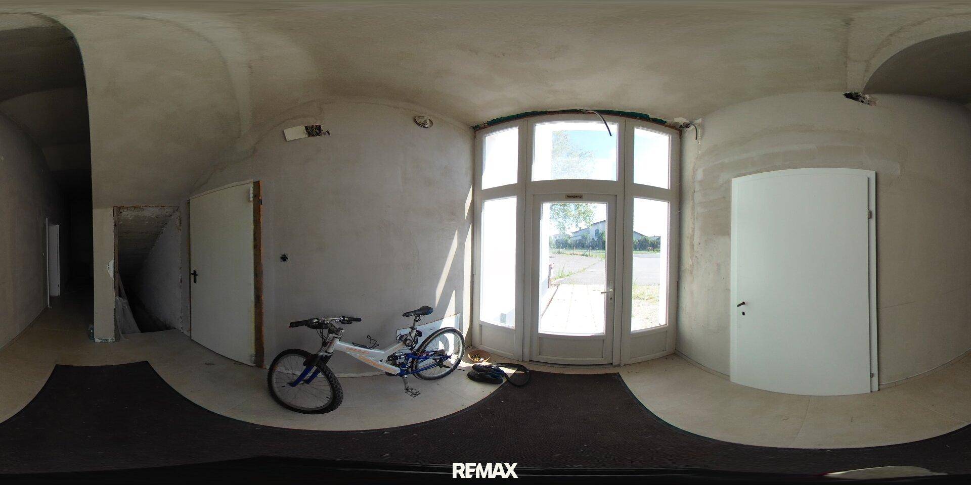 Eingangsbereich im Rohbau mit großer Glastür und unfertigen Wänden, ein Fahrrad steht daneben.