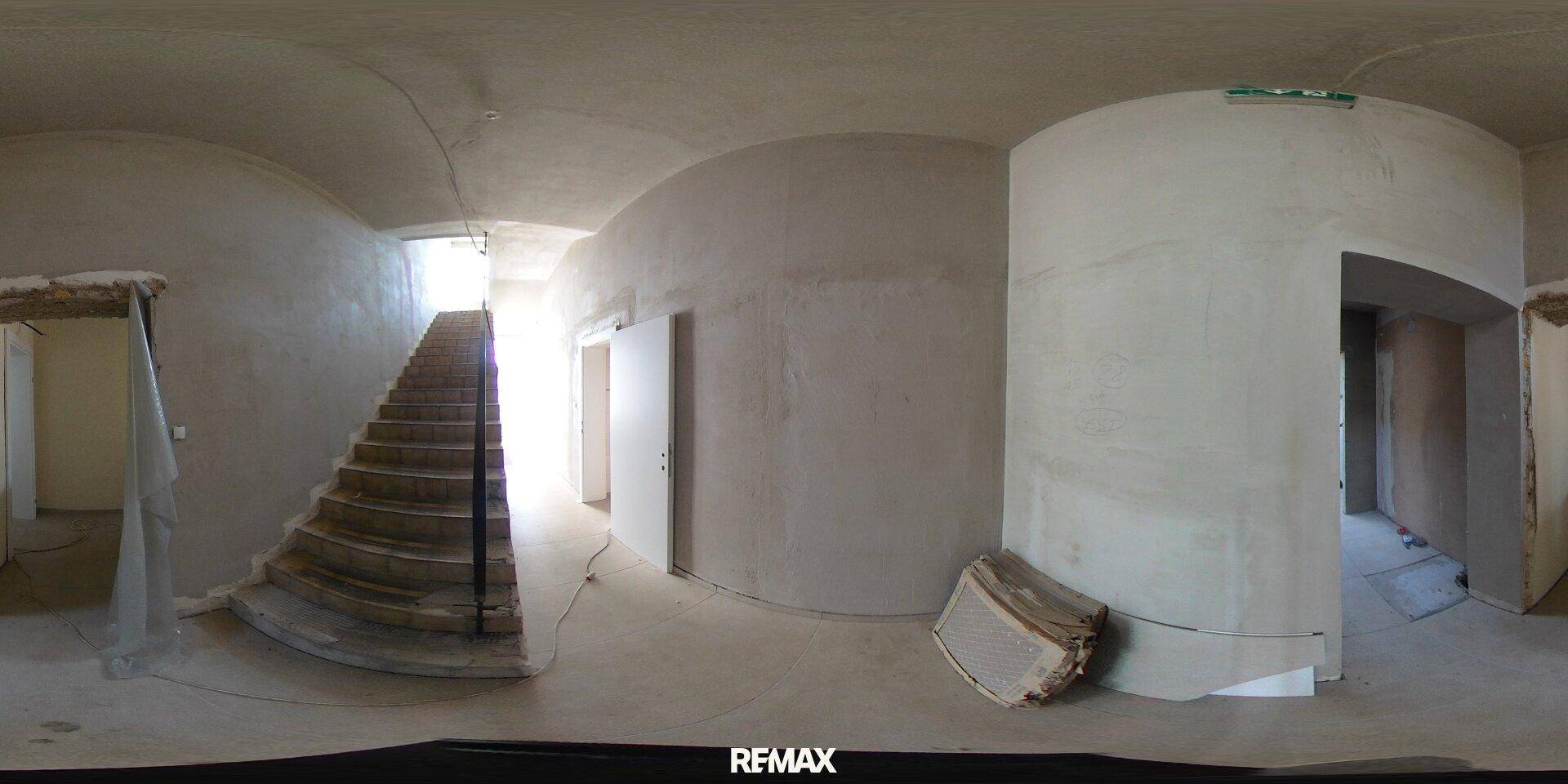 Flur im Rohbau mit einer geschwungenen Treppe, die in ein höheres Stockwerk führt.