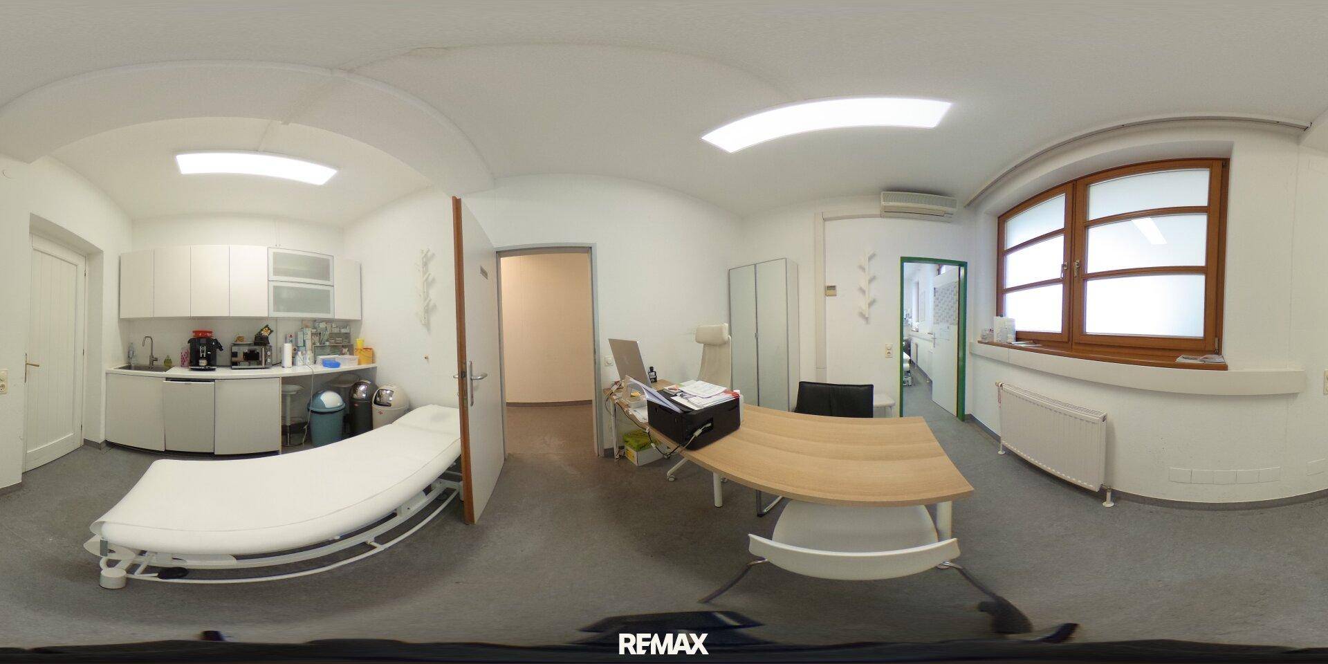 Panoramaansicht eines Untersuchungszimmers mit Küchenzeile, Schreibtisch und Behandlungsliege, praktisch eingerichtet.