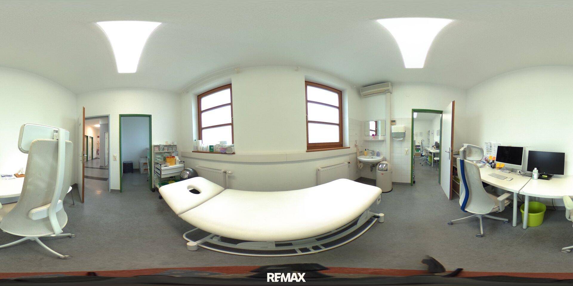 Panoramaansicht eines Untersuchungszimmers mit Behandlungsliege, Schreibtisch und Fenstern, gut beleuchtet und sauber.