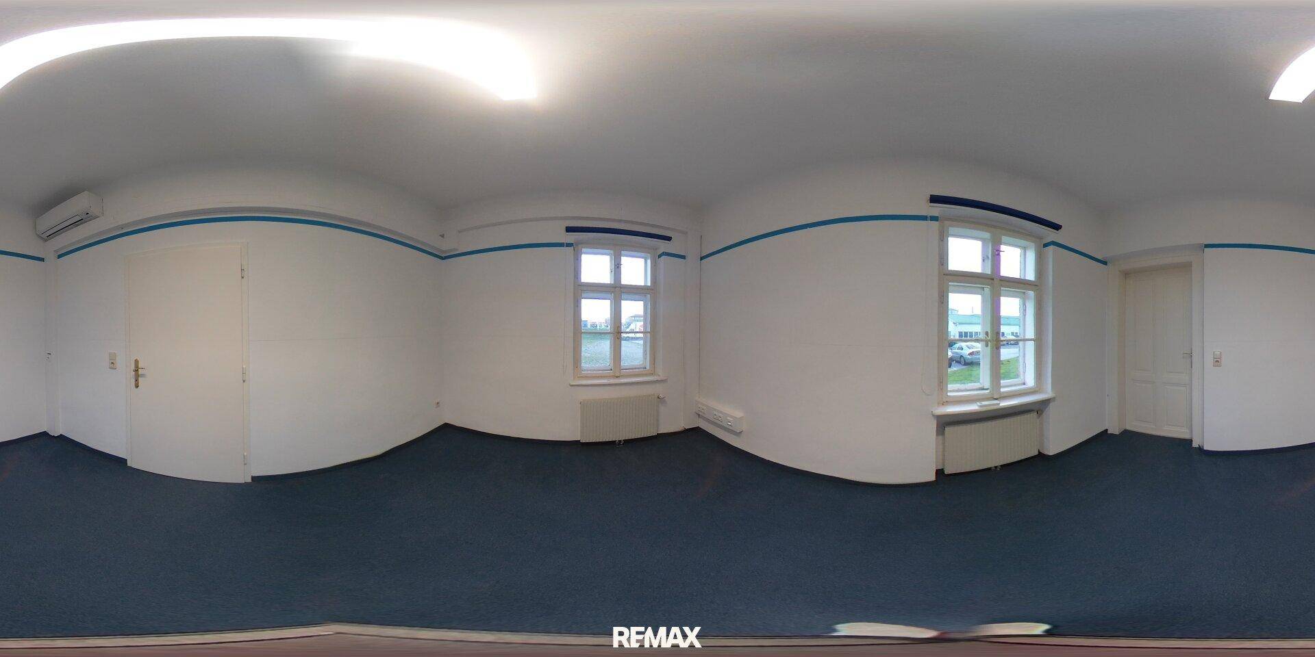 Panoramablick auf ein Büro mit blauem Teppichboden, weißen Wänden und zwei Fenstern.
