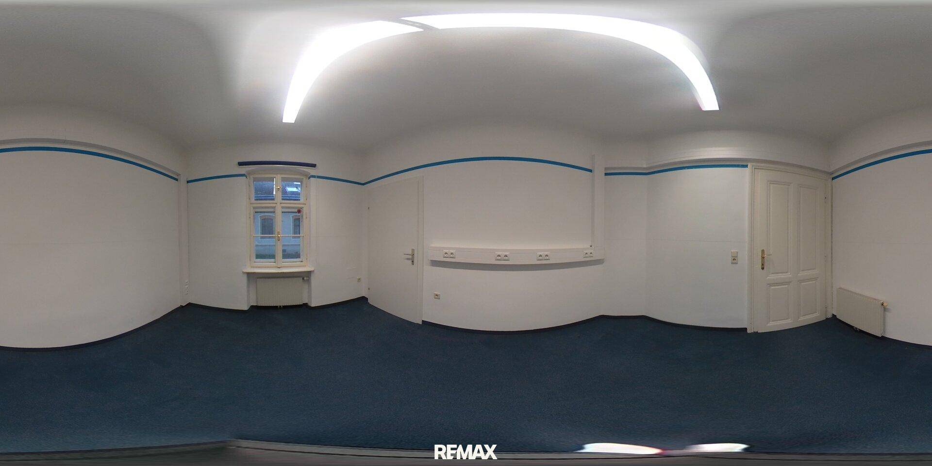Panoramablick auf ein Büro mit blauem Teppichboden, weißen Wänden und einem Fenster mit Heizkörper.