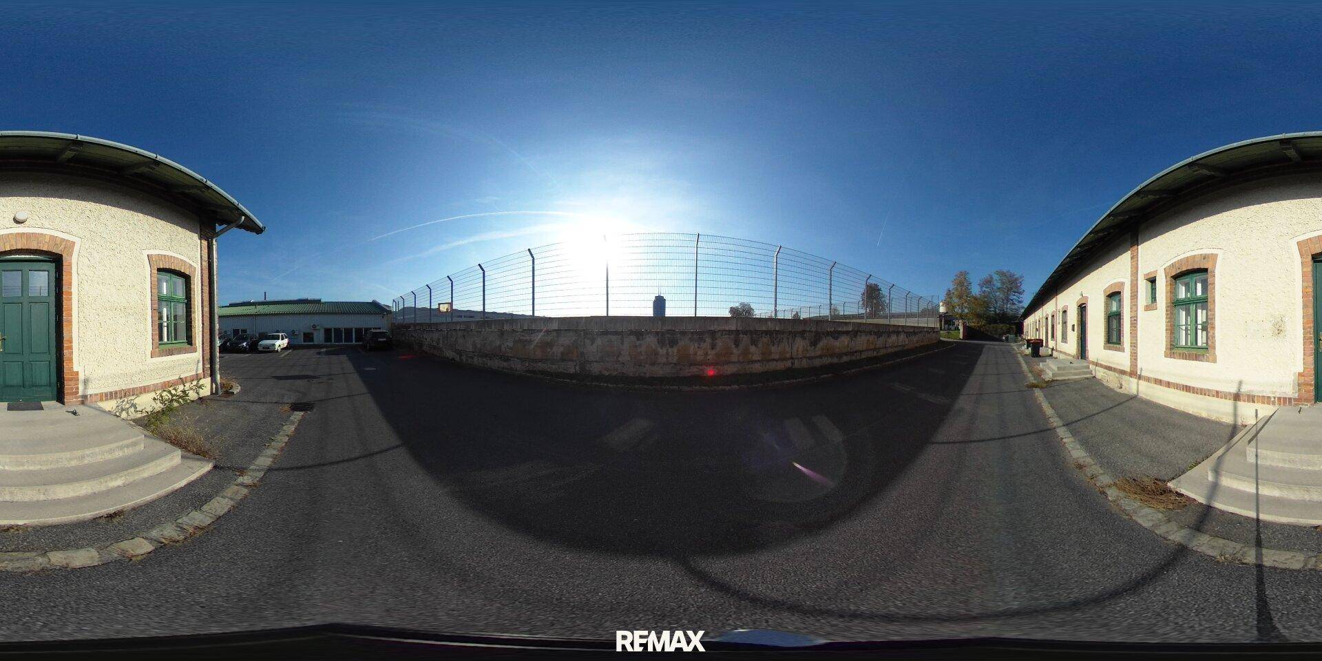 Panoramaansicht des Außenbereichs mit zwei Gebäuden, einem Zaun und blauem Himmel.