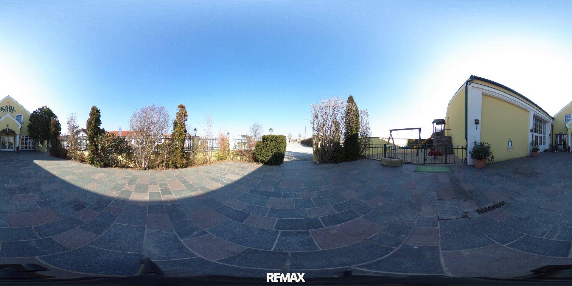 Panoramablick auf den gepflasterten Innenhof mit Bäumen, Sträuchern und dem Gebäude im Hintergrund.