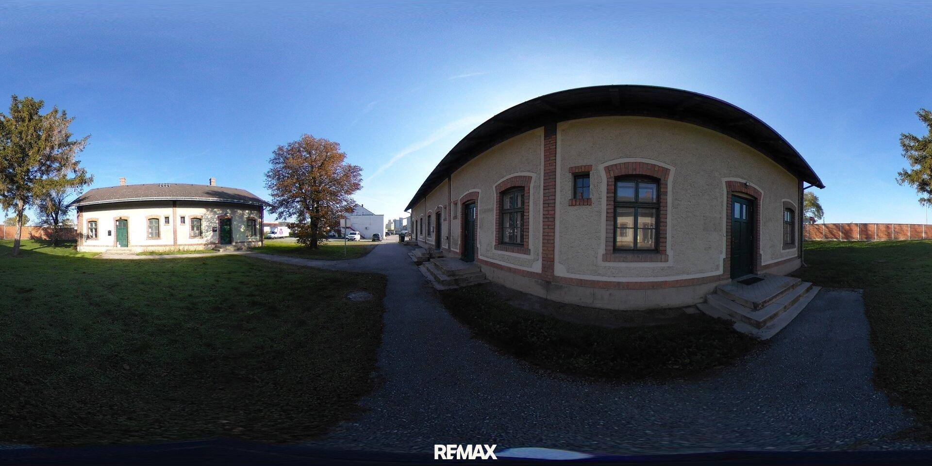 360-Grad-Außenansicht des Gebäudes mit umliegender Grünfläche und einem weiteren Gebäude im Hintergrund.