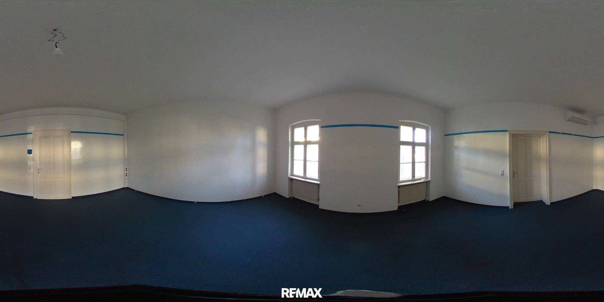 Weiter Raum mit dunklem Teppichboden, zwei Fenstern und Zugang zu weiteren Zimmern.