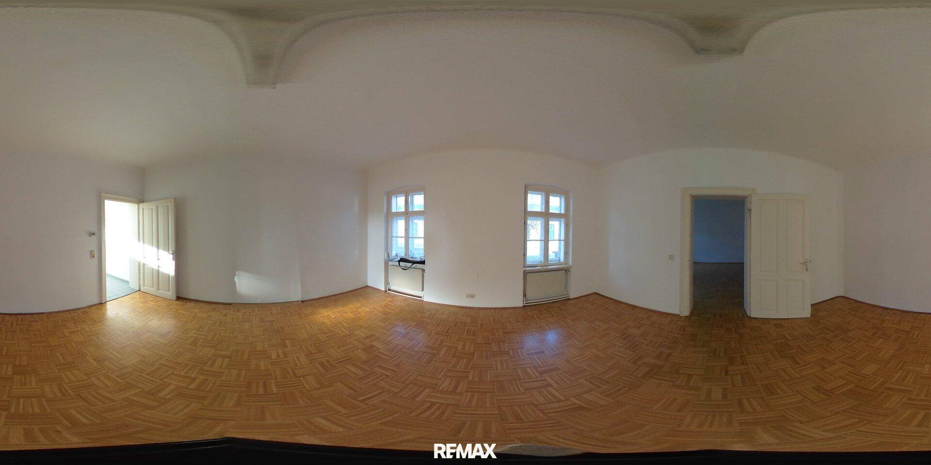 Leeres Zimmer mit Parkettboden und weißen Wänden, durch zwei Fenster fällt viel Tageslicht herein.