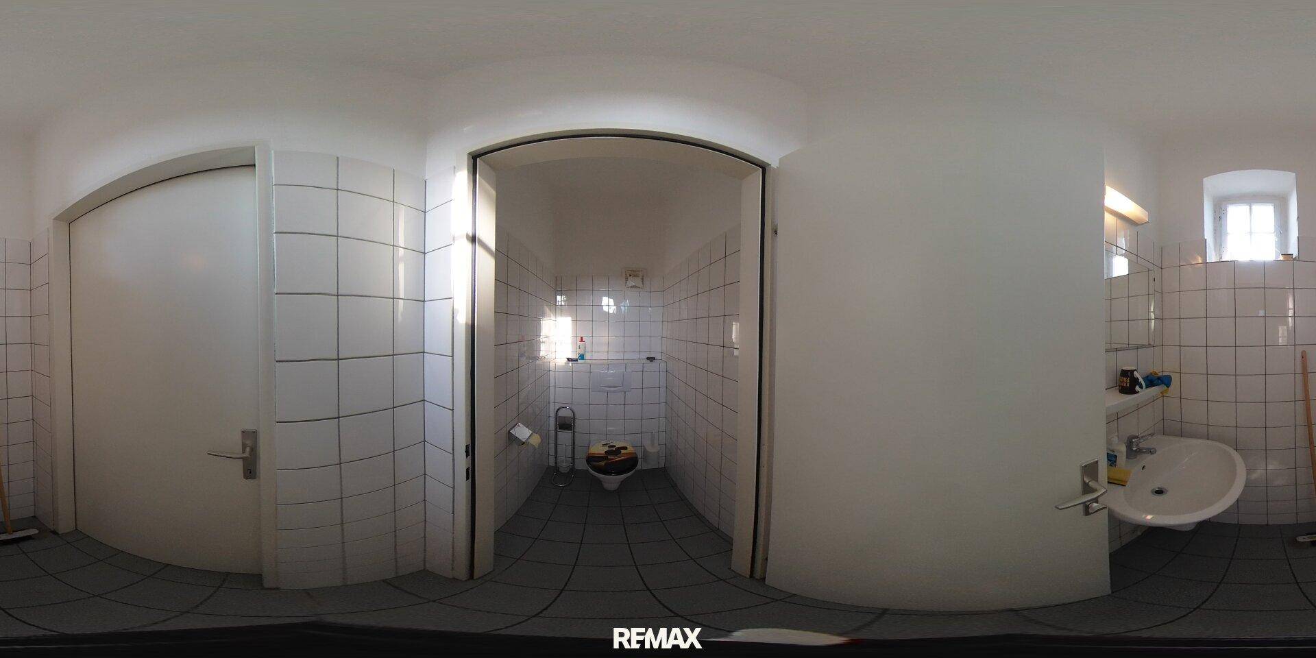 Badezimmer mit weißen Fliesen, Toilette und Waschbecken, durch ein kleines Fenster fällt Tageslicht herein.
