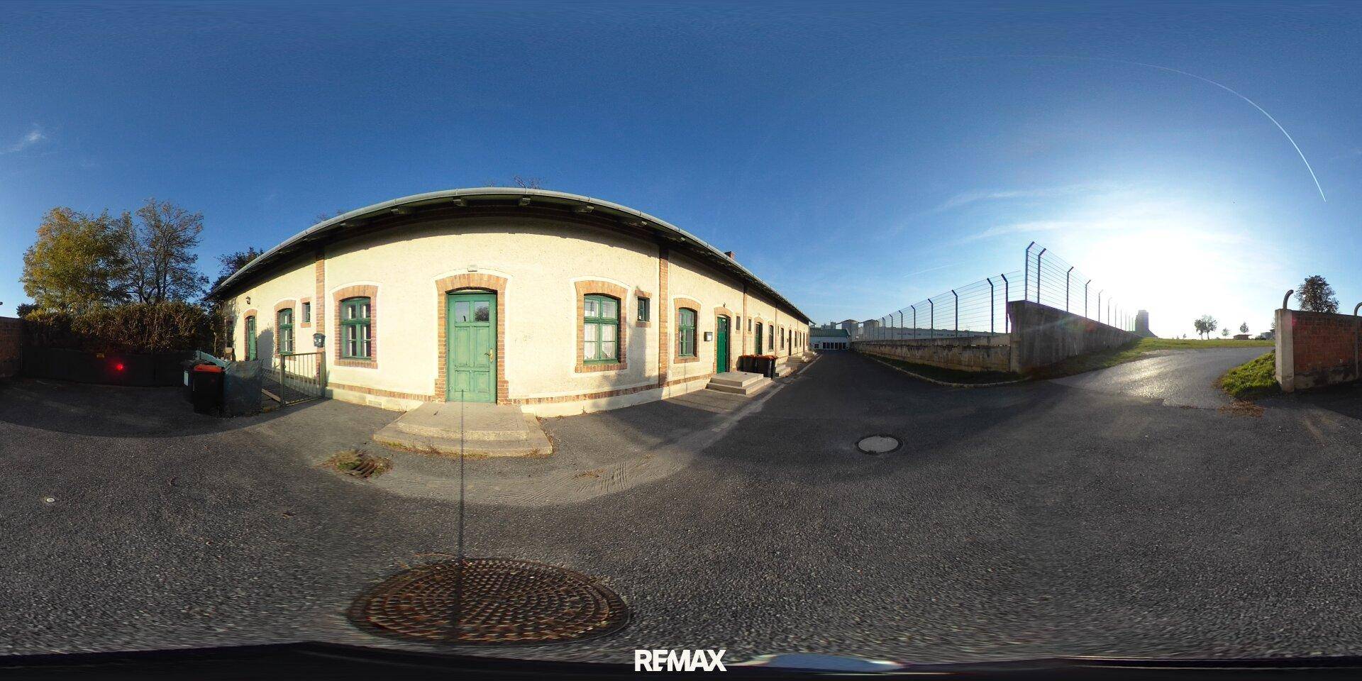 Panoramablick auf die Gebäudefassade mit asphaltiertem Vorplatz und einem Zaun unter blauem Himmel.