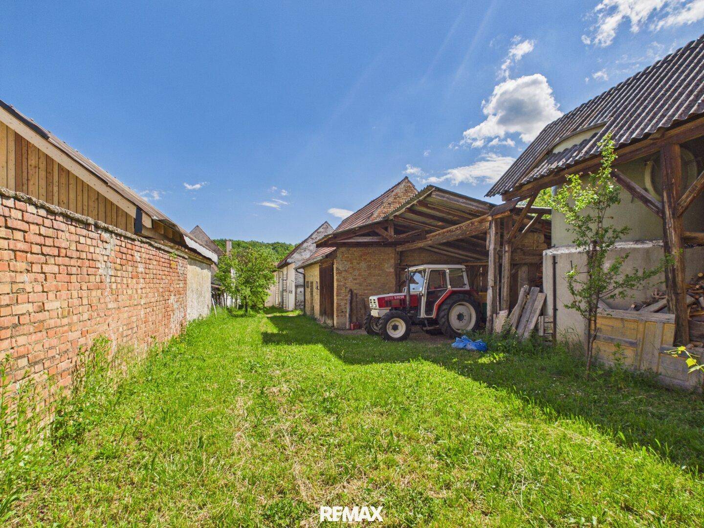 Langer, schmaler Grasweg zwischen Ziegelmauern und alten Wirtschaftsgebäuden mit Traktor.