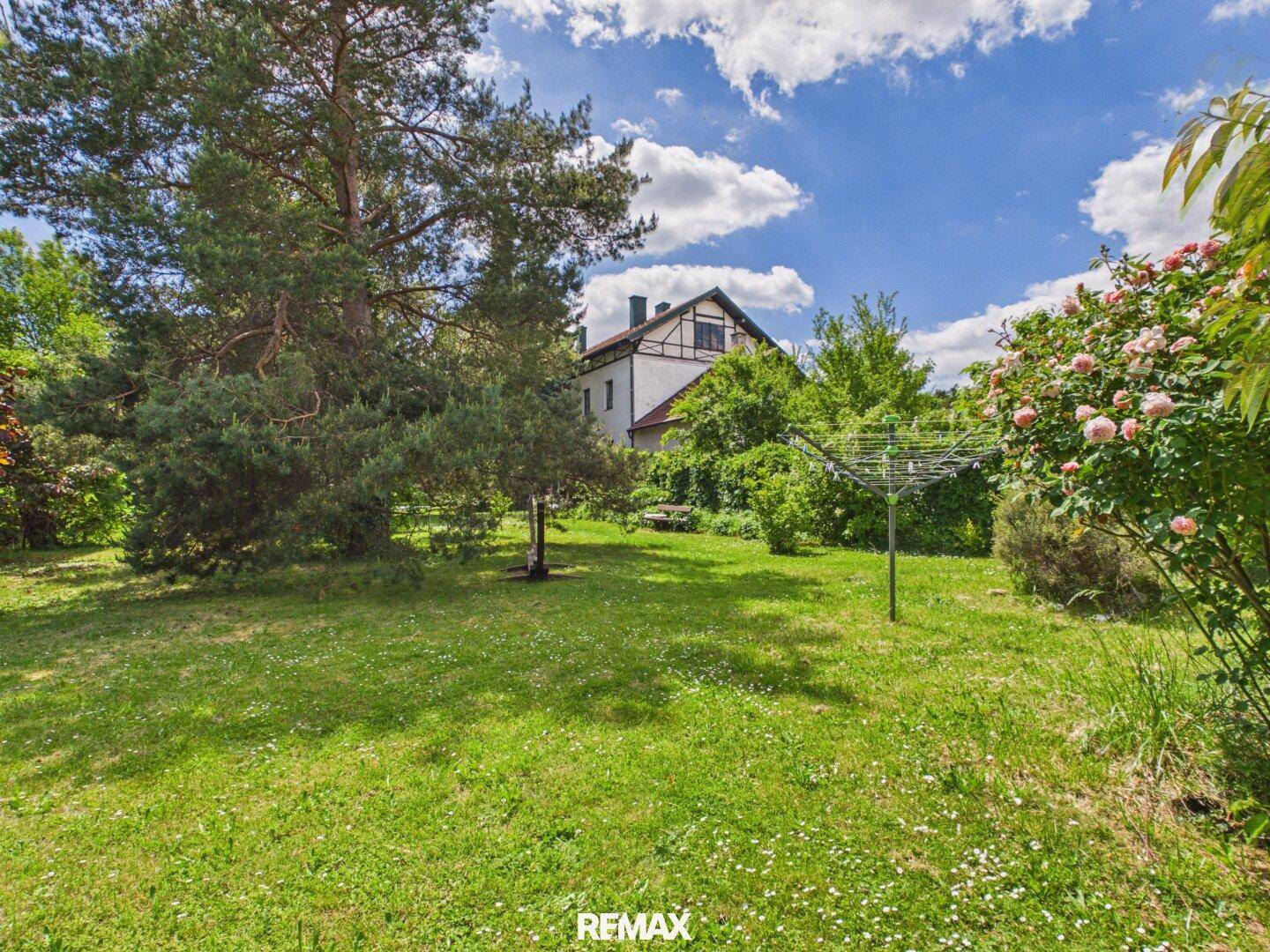 Weitläufiger Garten mit Rasen, hohen Bäumen und einem Wäscheständer, das Haus im Hintergrund.