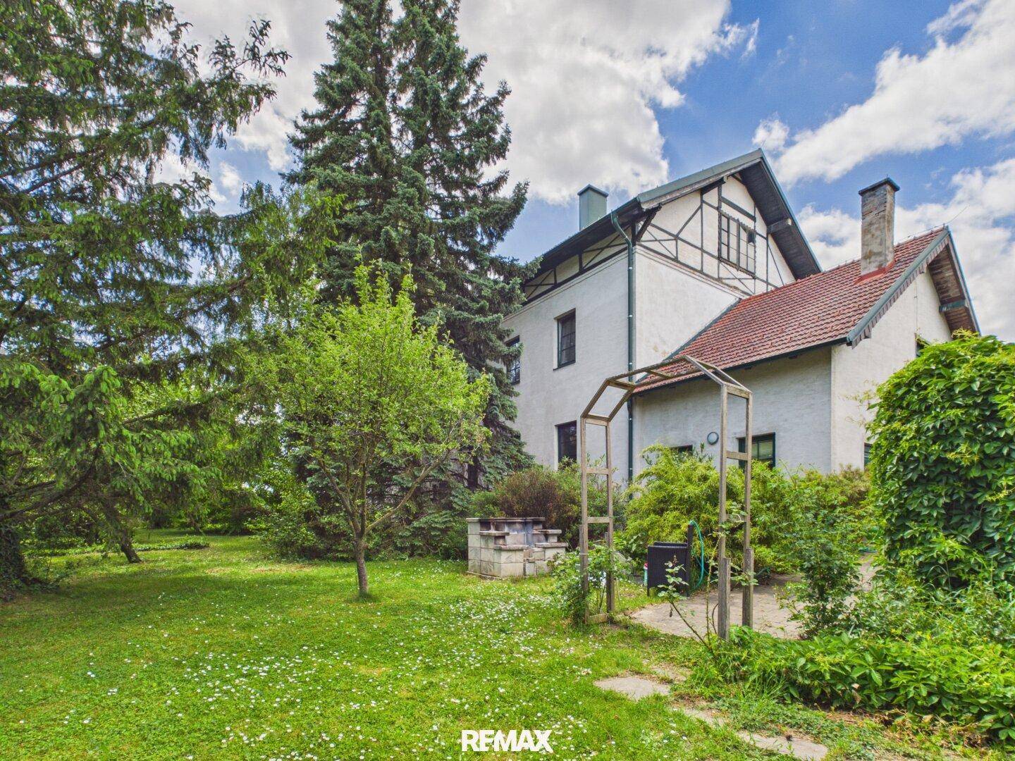 Großer Garten mit Rasenfläche, Bäumen und einem traditionellen Haus im Hintergrund unter blauem Himmel.