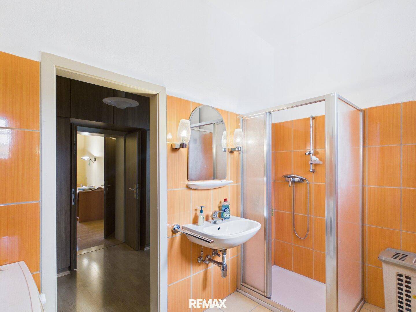 Badezimmer mit orangefarbenen Fliesen, Duschkabine und Waschbecken, das in einen anderen Raum führt.