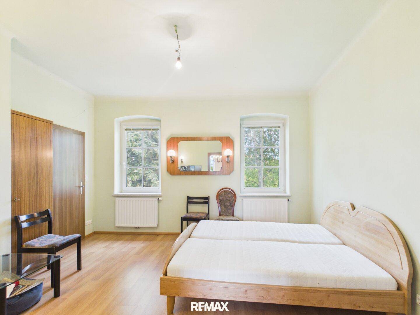 Helles Schlafzimmer mit Holzbett, Parkettboden und zwei Fenstern mit Blick ins Grüne.