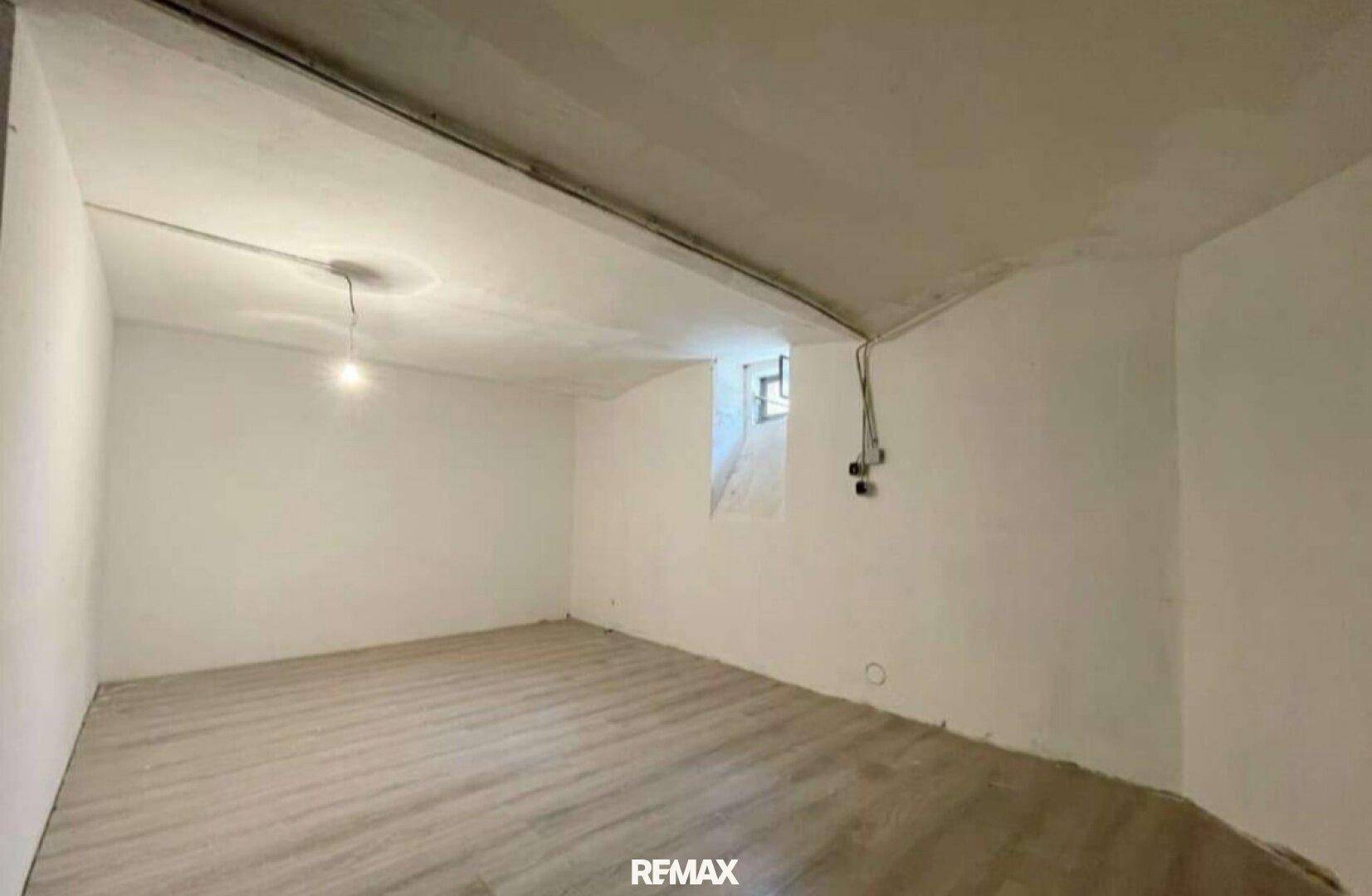 Unmöblierter Raum mit weiß gestrichenen Wänden, Laminatboden und einem kleinen Fenster, das Licht spendet.
