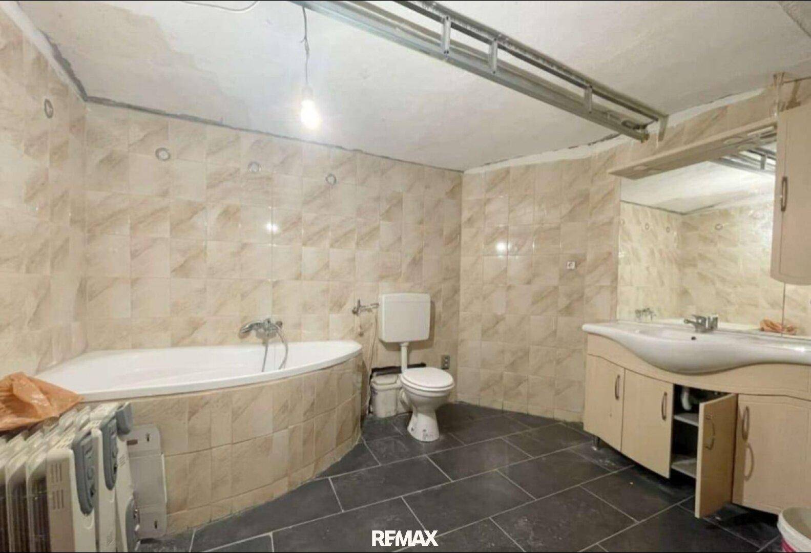 Badezimmer mit Eckbadewanne, WC und Waschtisch, ausgestattet mit beigefarbenen Wandfliesen und dunklem Fliesenboden.