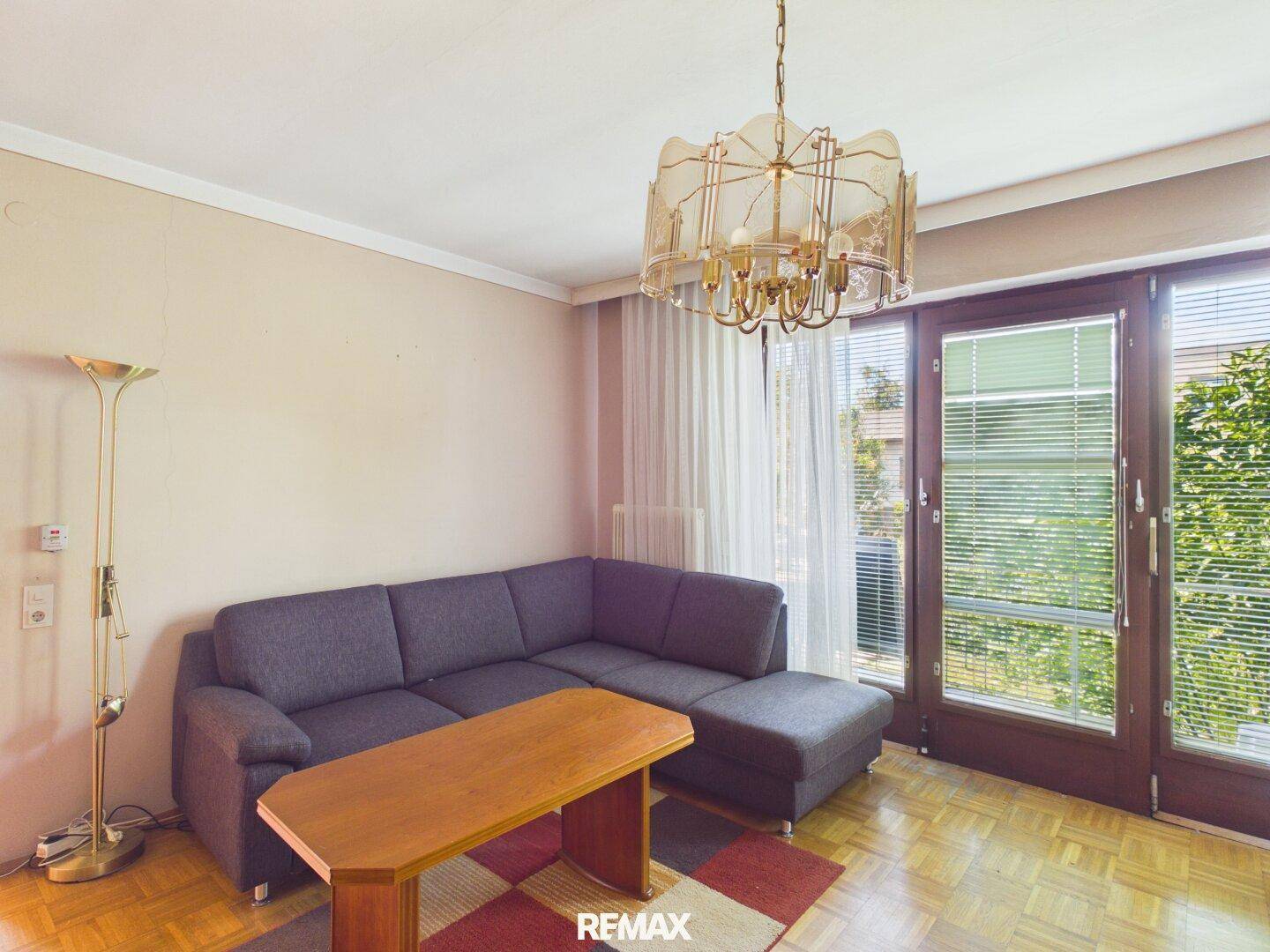Helles Wohnzimmer mit grauem Sofa, Holztisch und Zugang zu einem Balkon.