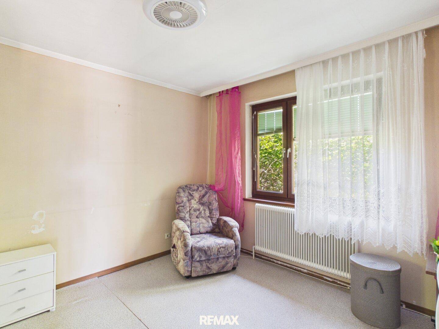 Schlafzimmer mit Teppichboden, einem Sessel und einem Fenster mit Blick ins Grüne.