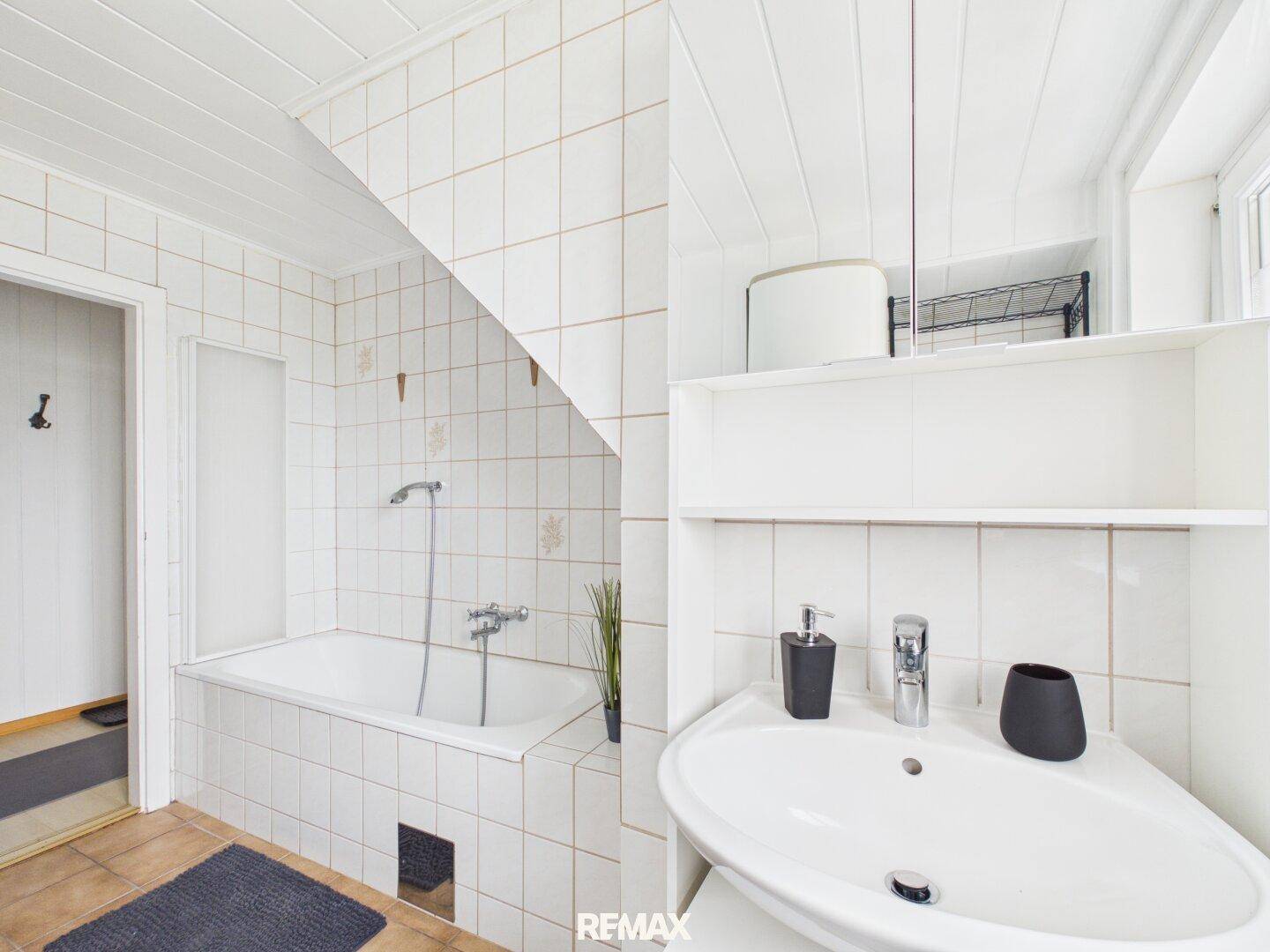 Helles Badezimmer mit weißen Fliesen, einer Badewanne mit Duschfunktion und einem Waschbecken unter der Dachschräge.